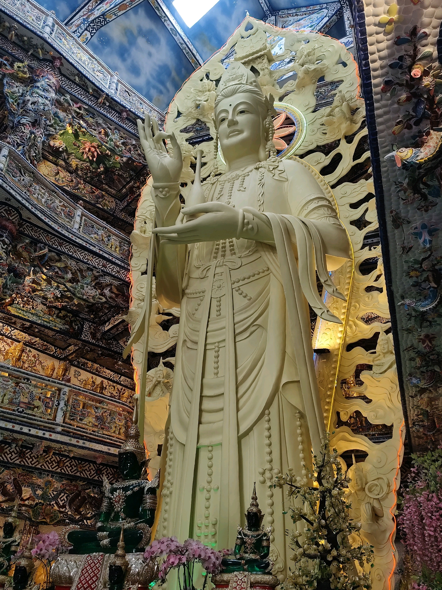 The image shows a large, white, standing statue of a deity, possibly Guanyin, in a temple setting in Xuân Thành, Vietnam. The statue is intricately detailed, with flowing robes and a serene expression. It is adorned with a crown and jewelry. The background is elaborately decorated with colorful mosaics and reliefs depicting various figures and scenes, characteristic of Vietnamese Buddhist temples. To the left of the main statue, there is a smaller, seated green statue of Buddha. In the foreground, there are decorative plants and flowers, adding to the spiritual ambiance of the location. The ceiling is visible in the upper part of the image, painted with a blue sky and clouds motif. The overall scene conveys a sense of reverence and artistic grandeur.