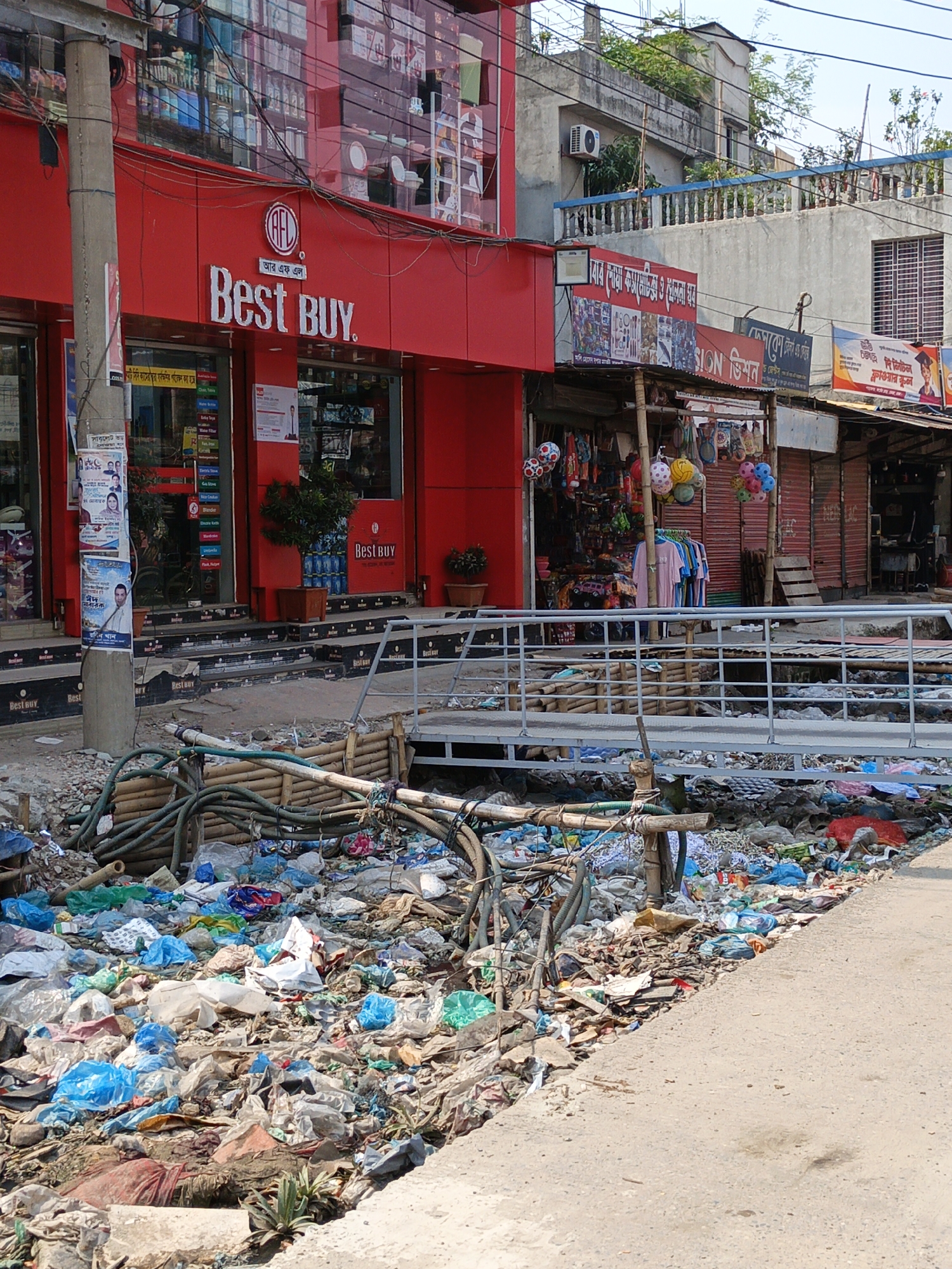 The image depicts a street scene in Dhaka, Bangladesh. A large amount of garbage fills a drainage ditch running alongside the road. The garbage includes plastic bags, discarded objects, and other debris. A makeshift bridge made of metal railings and wooden supports spans the ditch, allowing pedestrians to cross. Adjacent to the ditch and street, several businesses are visible. One is a modern retail store with a red facade and signage reading "Best BUY." Next to it is a smaller shop selling miscellaneous items, including clothing and toys. A utility pole stands near the "Best BUY" store, covered with posters. Further down the street, there are other commercial establishments with signage in Bengali. The environment suggests a densely populated urban area with significant waste management challenges.