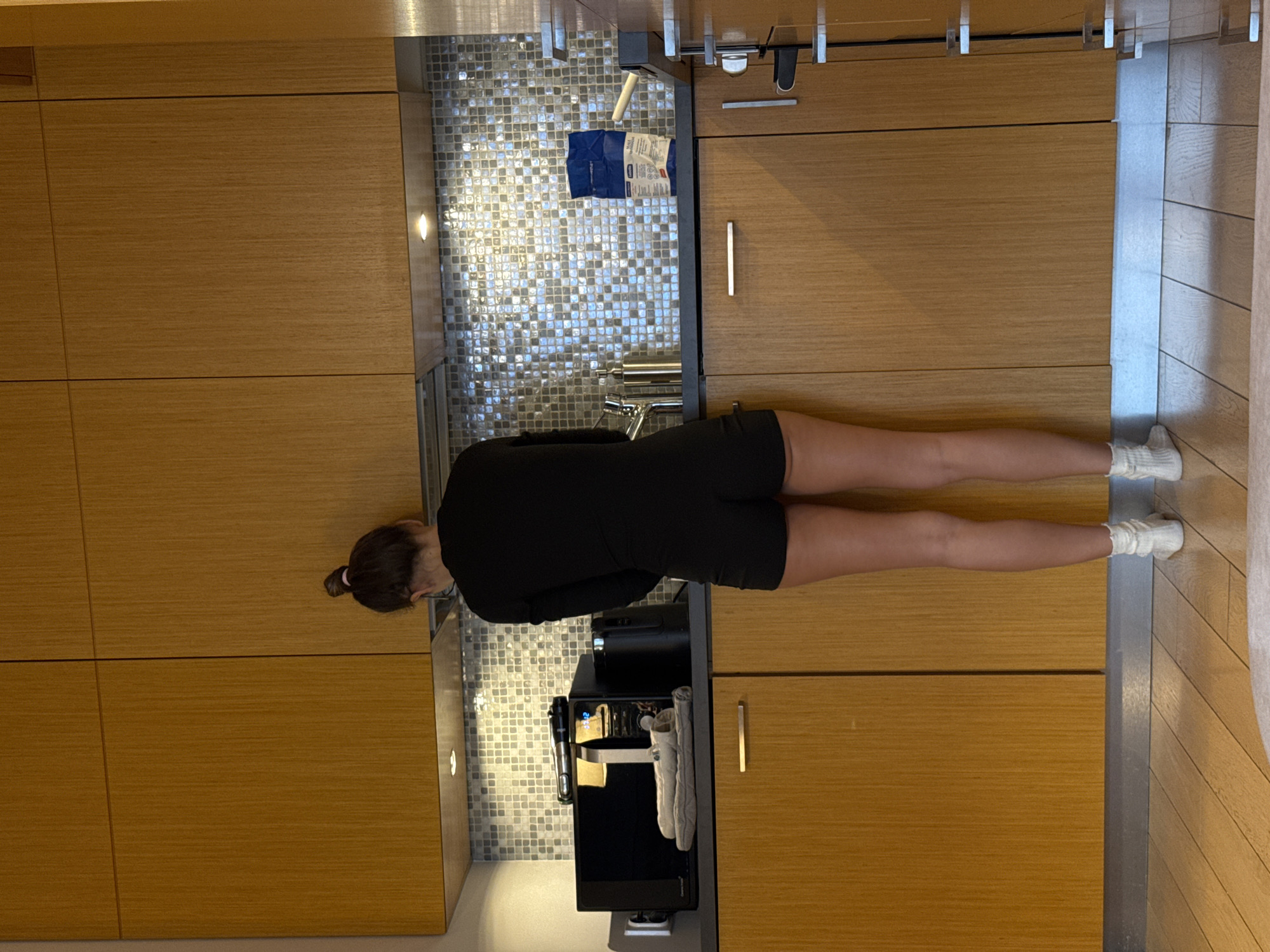 A person is seen from the waist up, positioned in a kitchen. They are wearing a black outfit and white socks. The kitchen features wooden cabinets, mosaic tile backsplash, and a coffee machine on the counter. The location is Warsaw, Poland.