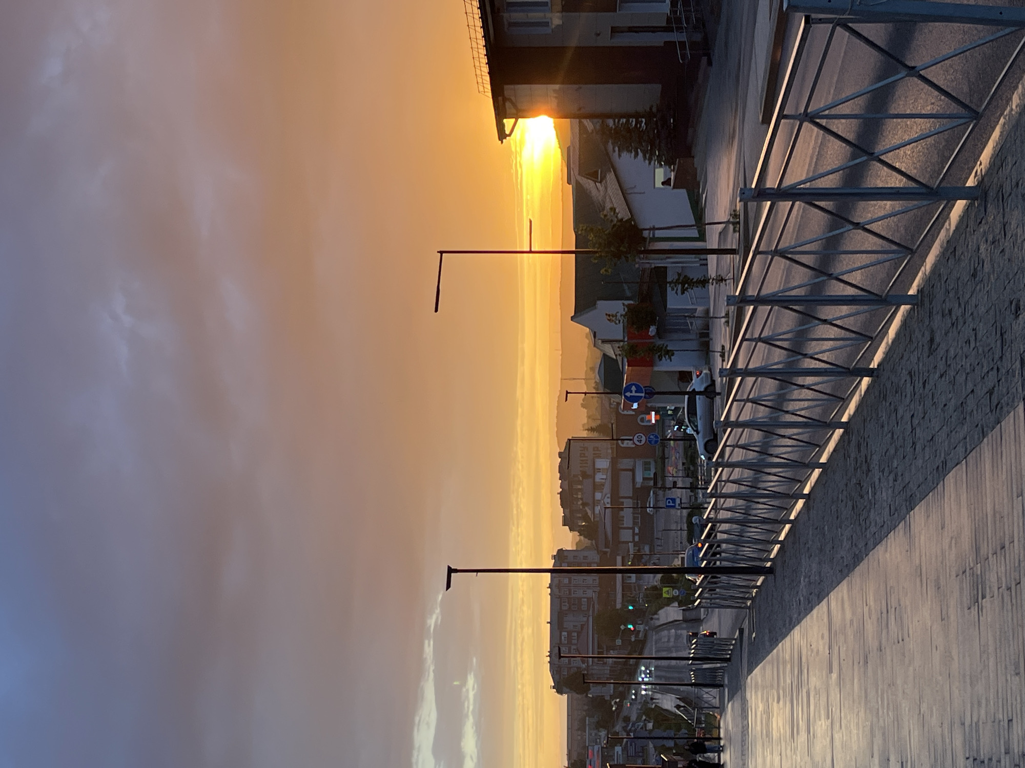 The image captures a scenic view of Kukmor, Russia, at sunset. A wooden walkway with a stone border stretches into the foreground, casting long shadows due to the low angle of the sun. A metal railing runs alongside the walkway, separating it from a road. The road features streetlights and is lined with buildings of varying architectural styles, indicative of a residential or commercial area within the city. Several cars are visible on the road, suggesting moderate traffic. In the background, more buildings and hills are dimly silhouetted against the vibrant orange and yellow hues of the sunset sky, which is partially obscured by clouds. The overall atmosphere is tranquil and serene, typical of an evening in a Russian town.