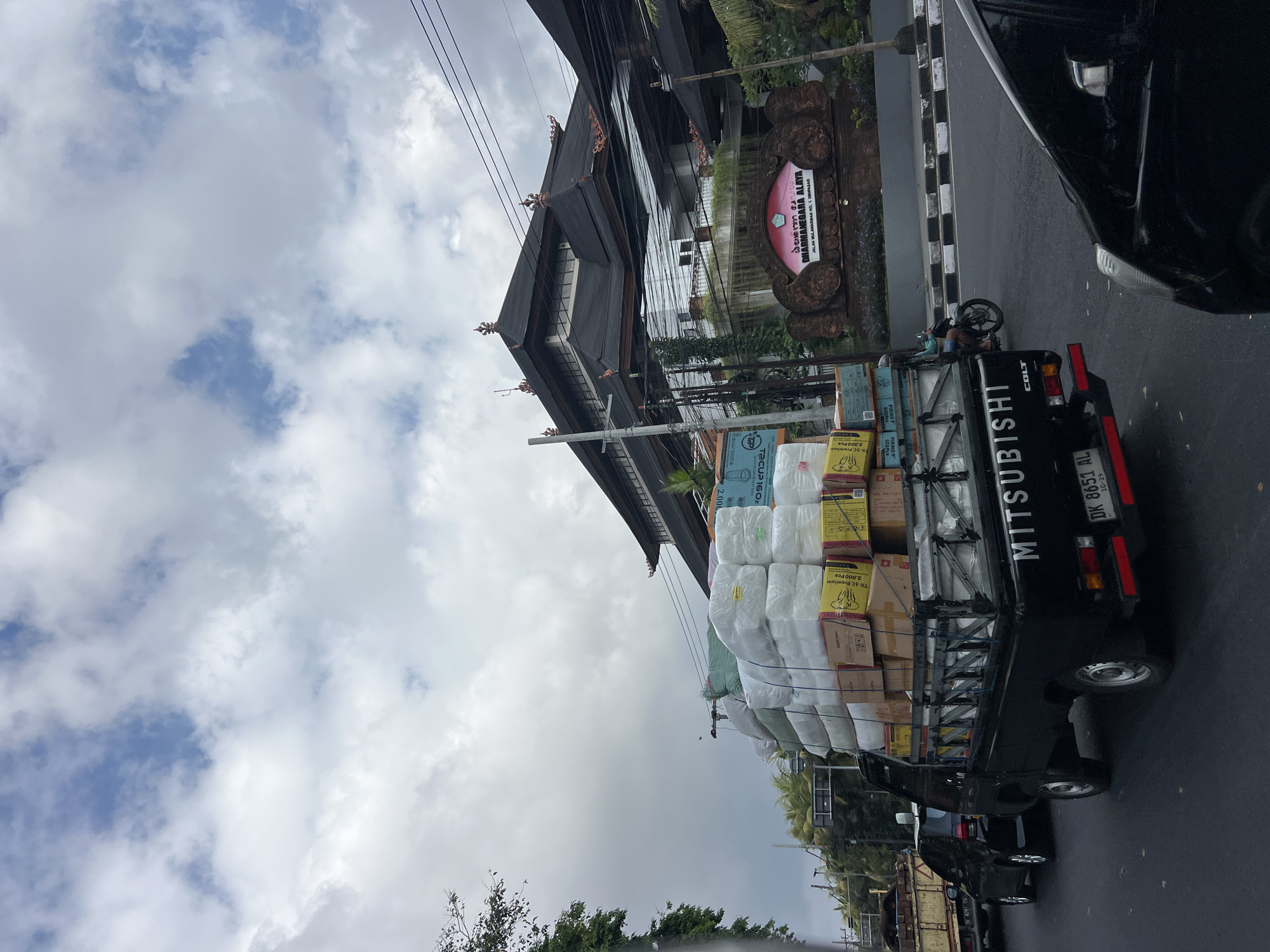 A black Mitsubishi Colt pickup truck is heavily loaded with boxes and white fabric bales, stopped at a street intersection in Denpasar, Indonesia. The truck displays the license plate DK 8651 AL. In the background, a large building with a traditional Balinese roof architecture is visible, with a sign that reads "DHARMANEGARA ALAYA JALAN MAHARADJAL NO. 1 DENPASAR." A motorcyclist is seen passing by the truck. The sky is overcast with scattered clouds. Other vehicles, including a black car and a utility truck, are also present at the intersection. Lush green trees and vegetation frame the scene.