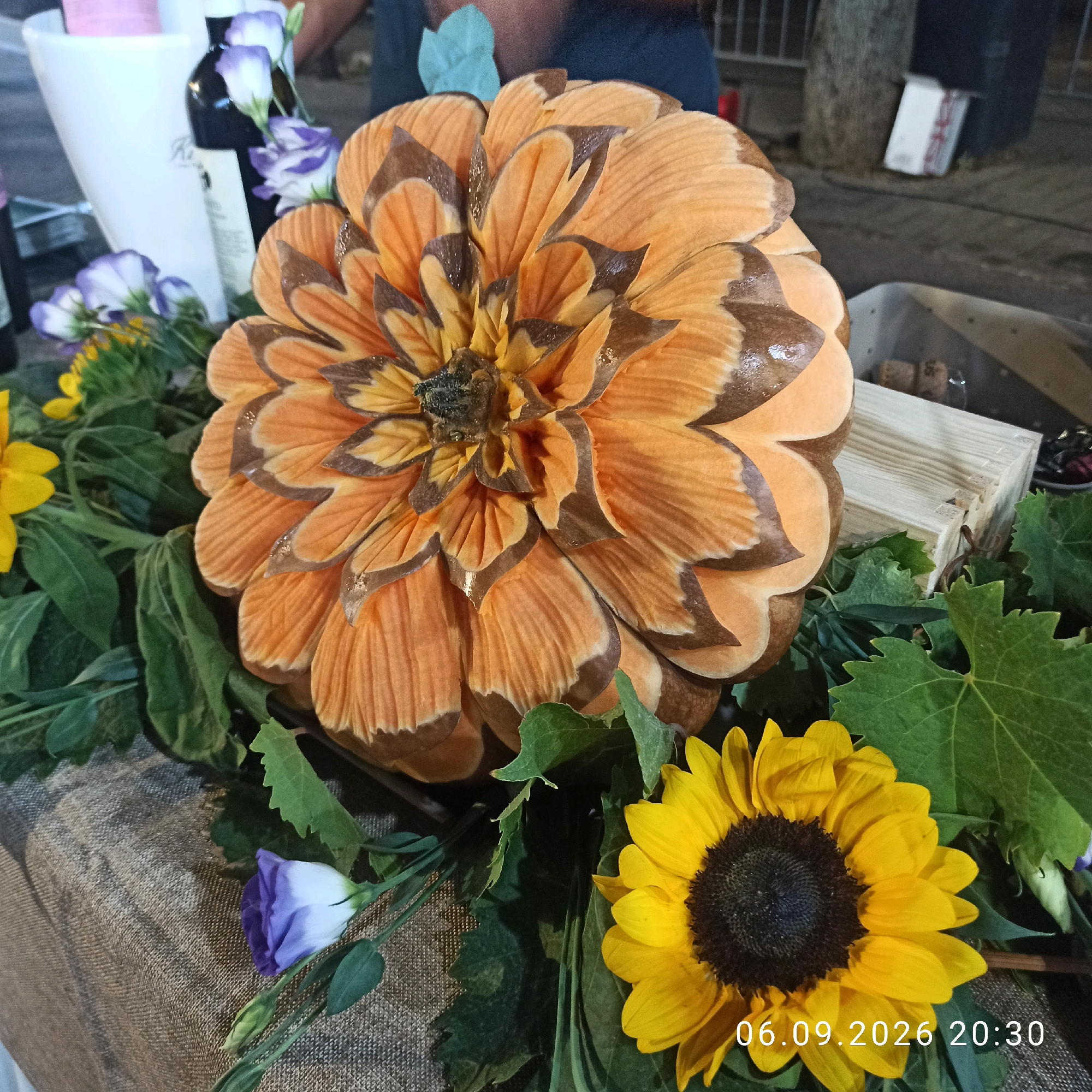 This is a close-up, eye-level shot of a table displaying floral arrangements and decorative items. The main subject is a large, intricately carved object resembling an orange dahlia flower, with layered petals in shades of orange and brown, and a dark center. Surrounding the dahlia are various flowers, including sunflowers with bright yellow petals and dark centers, and delicate purple and white lisianthus. Green foliage, including large grape leaves, fills the spaces between the flowers. The table is covered with a textured, burlap-like fabric. In the background, parts of bottles, a white bucket, and a wooden crate are visible, suggesting a market stall or event setting. The lighting suggests it is evening. A date and time stamp, "06.09.2026 20:30," is visible in the lower right corner. There are no people clearly identifiable in the frame, though the blurred figures in the upper background suggest human presence. The specific location in Bitonto, Italy, is not explicitly provided by the image itself.