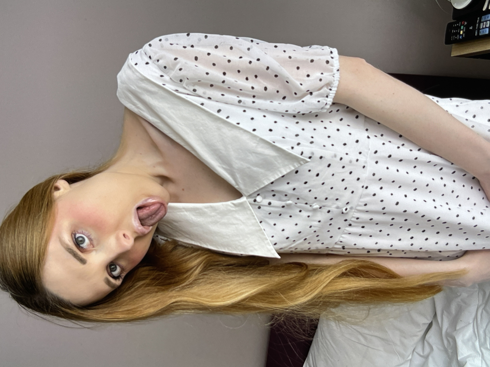 A young woman with long, blonde hair and blue eyes, wearing a white polka dot dress, is leaning forward with her tongue sticking out. She is indoors, in what appears to be a bedroom, with a white pillow visible on the left and a gray wall in the background. A remote control is visible on a wooden surface on the right.
