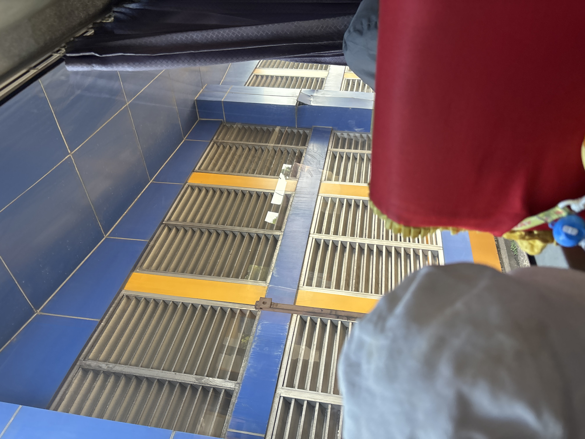 The image shows a view from inside a vehicle, looking out at a building with horizontal louvers. The building's facade is a combination of blue and orange tiles. A dark blue curtain is visible to the right, suggesting the vehicle is a bus or a similar mode of transport. In the foreground, parts of seats covered in red fabric and a person wearing a white head covering are visible. The scene is indoors, likely within the city of Purwakarta, Indonesia.