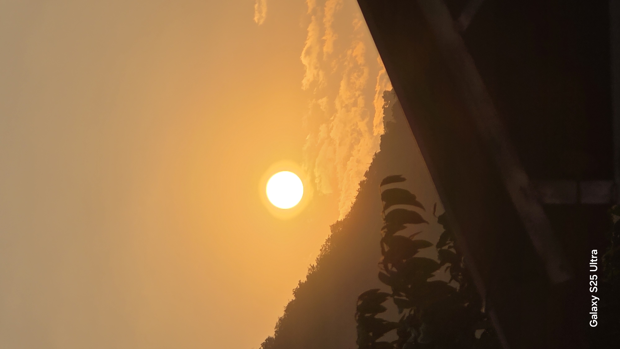 The image depicts a sunrise or sunset in the Bac Natural Heritage Area Trang, Vietnam. The sun is a bright, circular orb positioned in the upper-left quadrant of the image. There are sparse clouds near the sun. A silhouetted mountain and plant life occupy the lower-right portion of the image. Part of a structure is on the right, in the foreground, partially obstructing the view. The sky ranges in color from golden near the sun to a lighter, orange hue in the upper-left quadrant. "Galaxy S25 Ultra" is on the bottom right of the image.