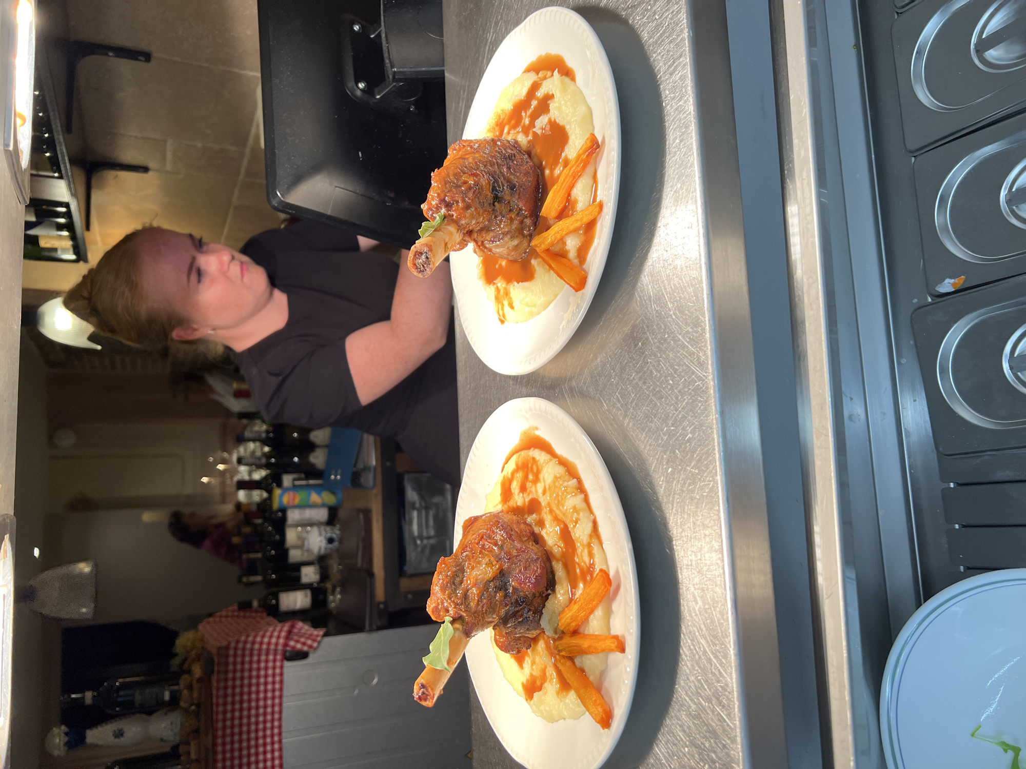 The image shows a close-up of two plates of food, likely in a restaurant kitchen or service area. The food appears to be lamb shanks served on a bed of mashed potatoes with a red sauce and garnished with carrot sticks and a sprig of green herb. The plates are white and have a textured rim.
In the background, a woman with red hair pulled into a bun is visible, wearing a black t-shirt and looking at a computer monitor. Behind her, shelves are stocked with bottles of wine and other items. A red and white checkered cloth is draped over a counter on the left. The environment appears to be a professional kitchen setting with stainless steel surfaces.
The image does not provide any specific information or visual cues that would allow for its location to be identified as the city of Guildford, United Kingdom.