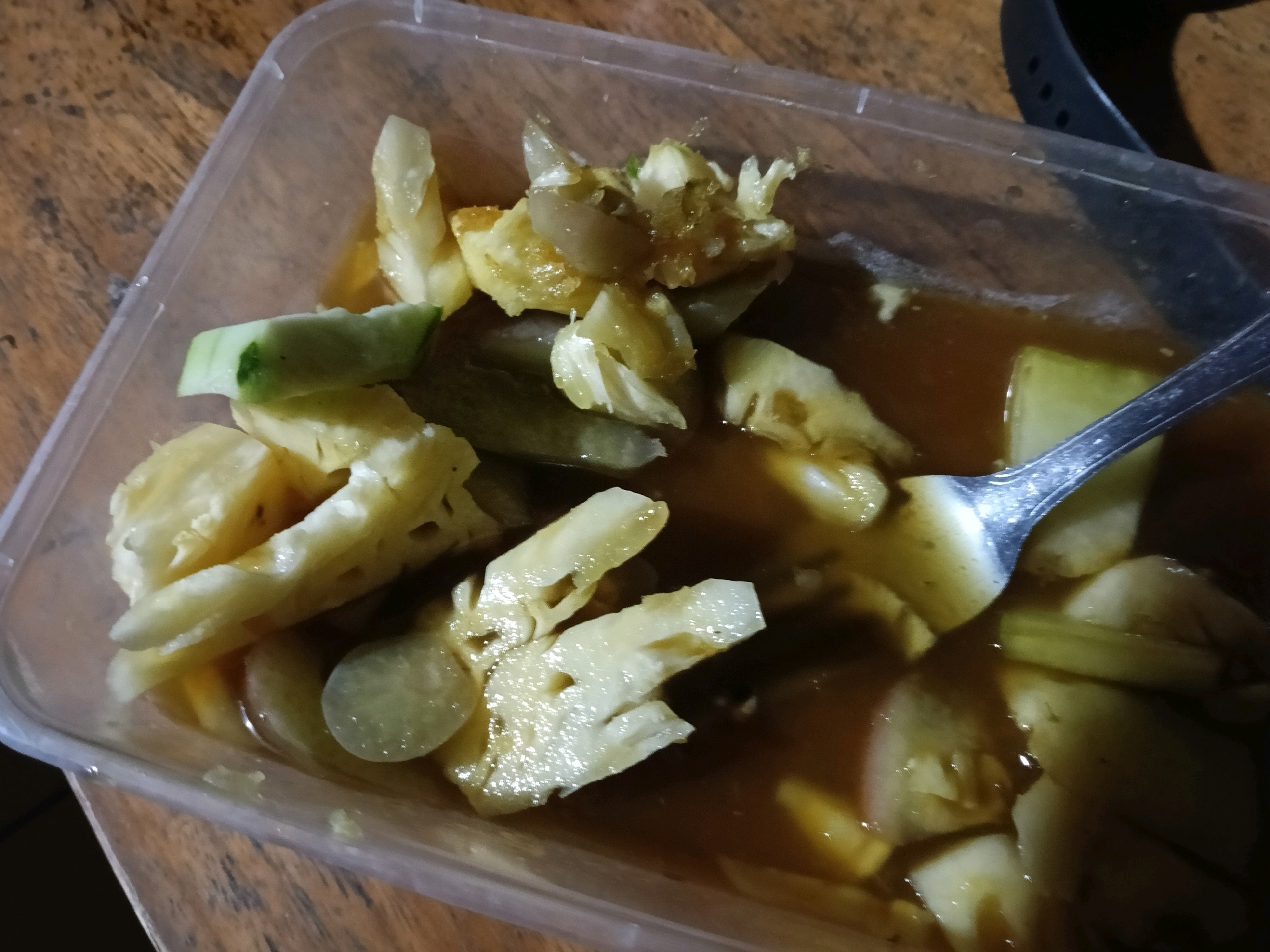 A clear plastic food container filled with a dish of pineapple and cucumber salad in a brown sauce is pictured on a wooden table. The container is positioned at an angle. A metal spoon is partially submerged in the salad. The background is blurred and appears to be a wooden surface. No people or other activities are visible. The location is identified as Galuk, Indonesia.