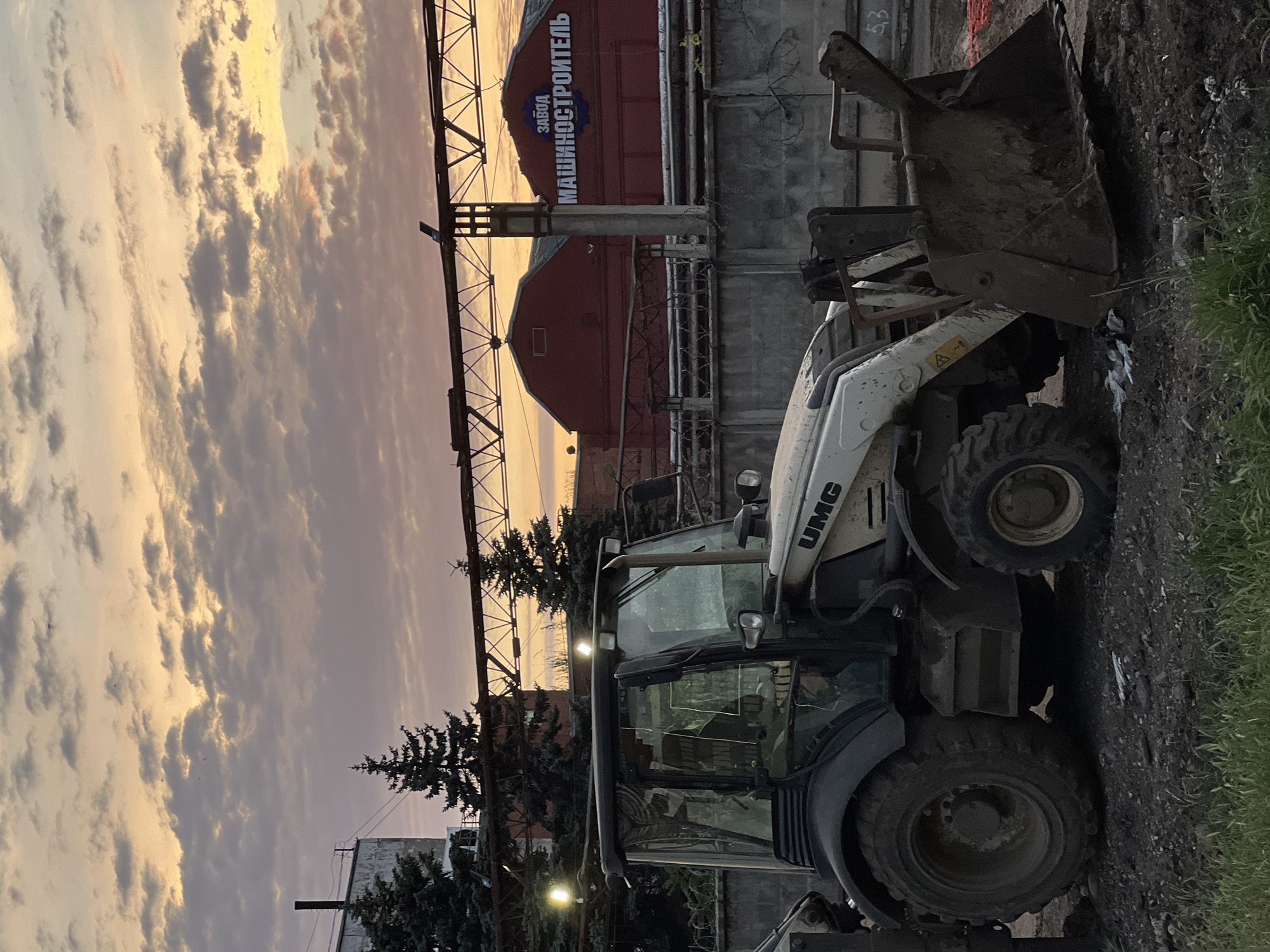 An outdoor, eye-level, medium shot captures a white and black UMG backhoe loader parked on a dirt surface in front of a concrete fence and industrial buildings. The backhoe loader faces right, with its front loader bucket lowered to the ground. Two bright lights are visible on the roof of the backhoe's cabin. Behind the backhoe, a corrugated metal fence runs horizontally, topped with barbed wire. In the background, two large, red industrial buildings stand, one with a blue circular logo and white Cyrillic text that reads "ЗАВОД МАШИНОСТРОИТЕЛЬ" (Manufacturer Plant). A metal truss structure extends across the top of the image, supporting overhead wires. Tall, dark green evergreen trees are situated to the left of the buildings. The sky above is filled with dramatic clouds in hues of orange, pink, and gray, indicating either sunrise or sunset. The ground is covered in dark soil, gravel, and some green grass in the foreground. The overall impression is an industrial or construction site during twilight.
