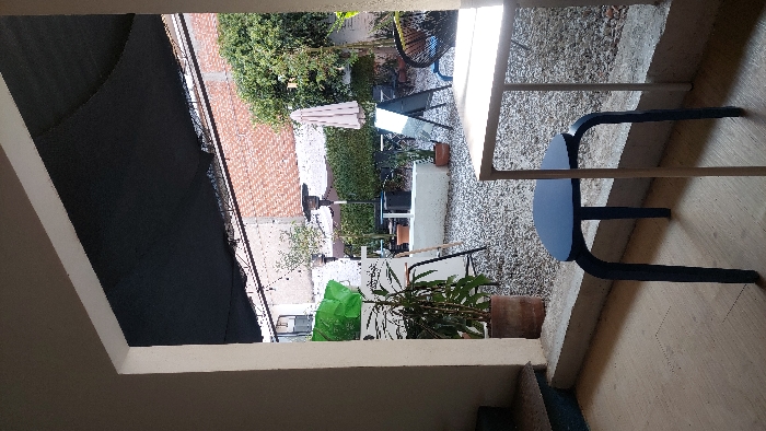 The image depicts an outdoor patio or courtyard viewed from an interior threshold. The foreground shows a light-colored floor, a dark blue four-legged stool, and the edge of a light-colored table. A concrete ledge separates this interior space from the outdoor area. The outdoor patio, located in Tlaxcala City, Mexico, is surfaced with light-colored gravel and partially covered by a dark mesh shade cloth supported by a metal frame.
Within the patio, a large potted plant in a terracotta pot is positioned on the left, alongside a white counter-like structure topped with green material and a black A-frame sign. A tall, dark patio heater stands further back. In the center, a low, light-colored concrete barrier or planter contains smaller potted plants. To the right, a folded light-colored market umbrella is visible, accompanied by another black A-frame sign and a black and blue woven chair. The background features a reddish-brown brick wall, with parts of a light-colored urban building and sparse green foliage visible above and behind it. No individuals are present in the scene.