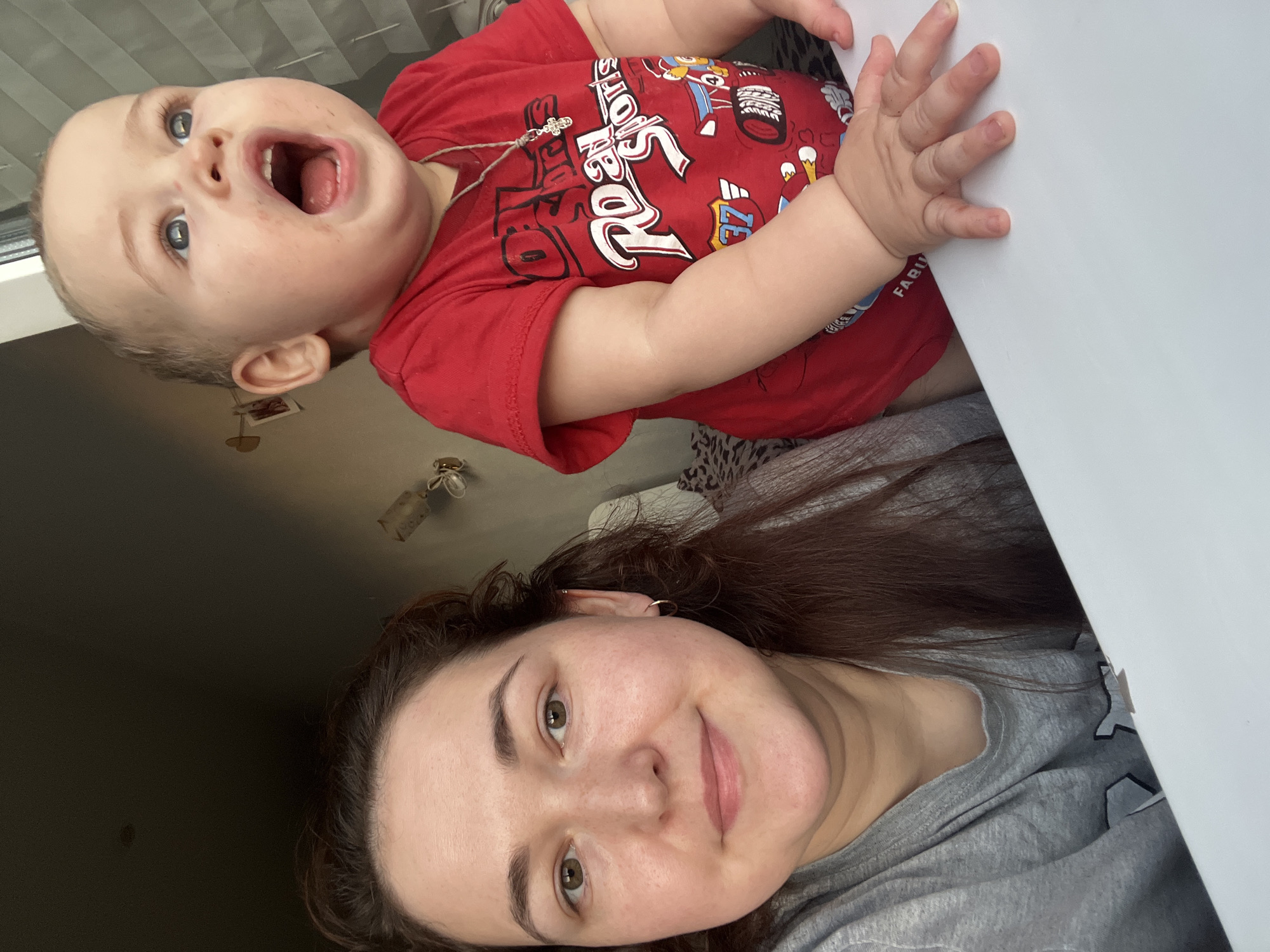 This media file is a close-up, tilted selfie of two people. In the foreground, a young woman with brown hair and brown eyes is smiling. She is wearing a grey t-shirt. In the background, a baby with blue eyes and an open mouth is wearing a red t-shirt with graphics and text. The baby's hand is visible, touching a white surface. The setting appears to be indoors, possibly a home. The lighting suggests it could be daytime. The overall mood is cheerful and playful. The text on the baby's shirt is partially visible and includes the words "ROCK" and "STARS". A small cross necklace is also visible on the baby.