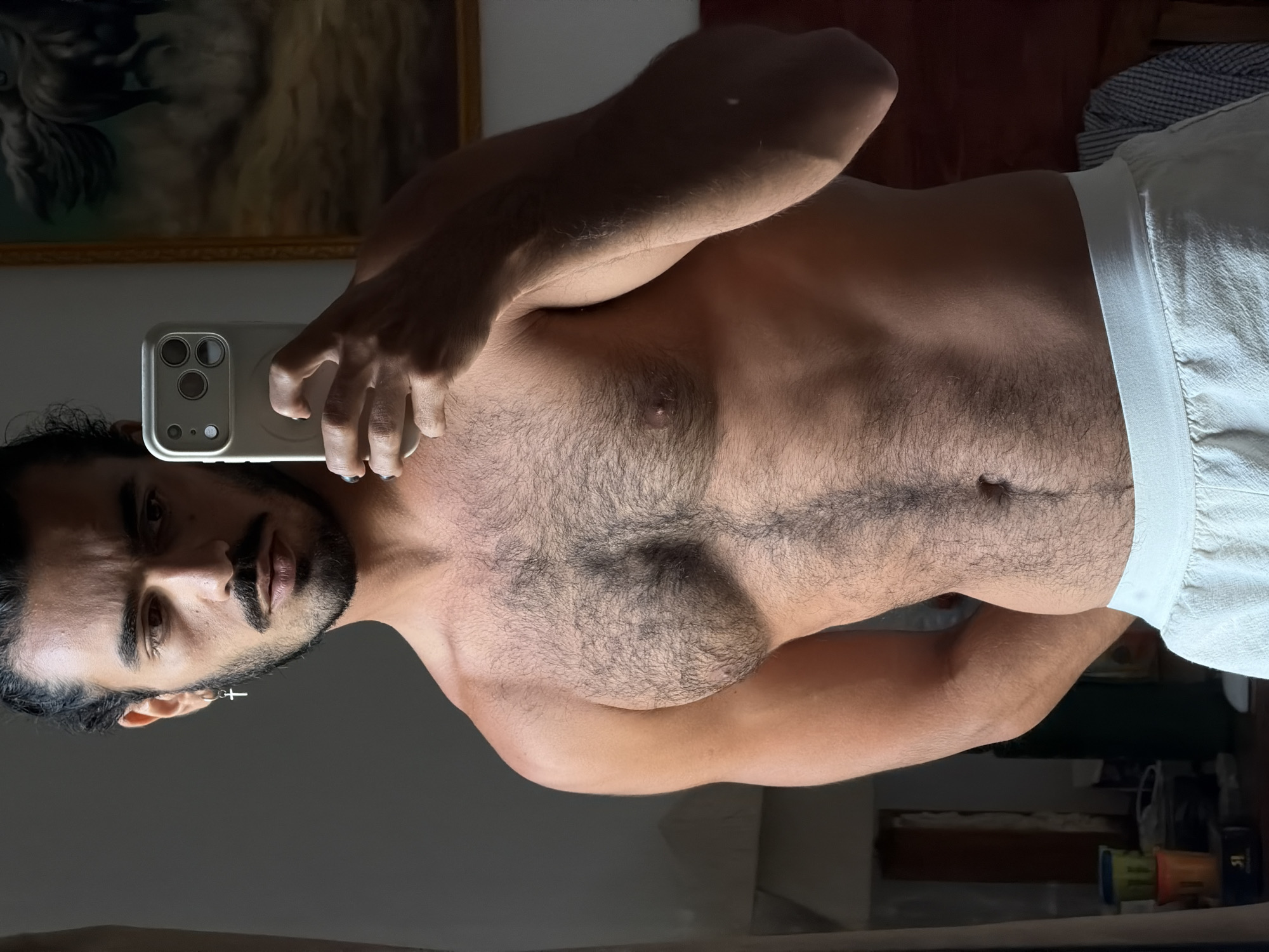 The image depicts a shirtless man taking a selfie in a dimly lit room, likely a bedroom, as evidenced by the presence of a painting on the wall and a bedside table with items. The man has dark hair tied back, a mustache, and a cross earring. He is wearing light-colored shorts. The room appears to be residential, with a painting of a horse in the background and a bedside table with various items. The overall lighting suggests it's either early morning or late evening. The exact location in North Kuta, Indonesia cannot be determined from the image.