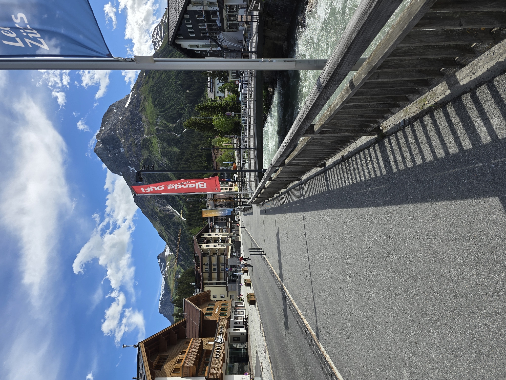 The image shows a street scene in Lech, Austria, on a sunny day with scattered clouds. A paved road runs through the center, bordered by a wooden railing on the right, overlooking a fast-flowing river. On the left side of the road, there are several traditional wooden buildings with balconies, typical of Alpine architecture. A prominent mountain with snow-capped peaks dominates the background. Two individuals are cycling on the road. Flags are visible, one red and one blue, with some text on them. The environment appears to be a town square or main street, with shops and residential buildings.