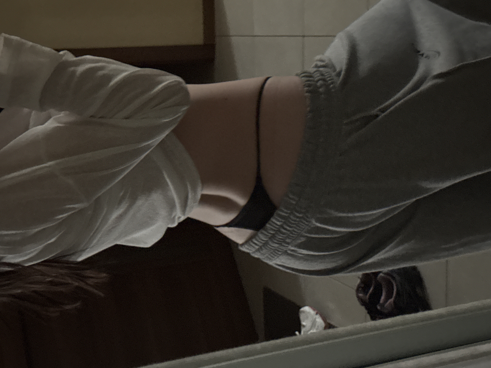 A person is partially visible in the image, shown from the waist down, wearing grey sweatpants and a white top pulled up to expose their midriff and a black thong. The person is standing in what appears to be a room with tiled flooring and a dark wooden door in the background. A mirror is partially visible on the left side of the image. The overall lighting is dim, suggesting it might be indoors at night. The image does not provide any information about the location in the city of Khimki, Russia.