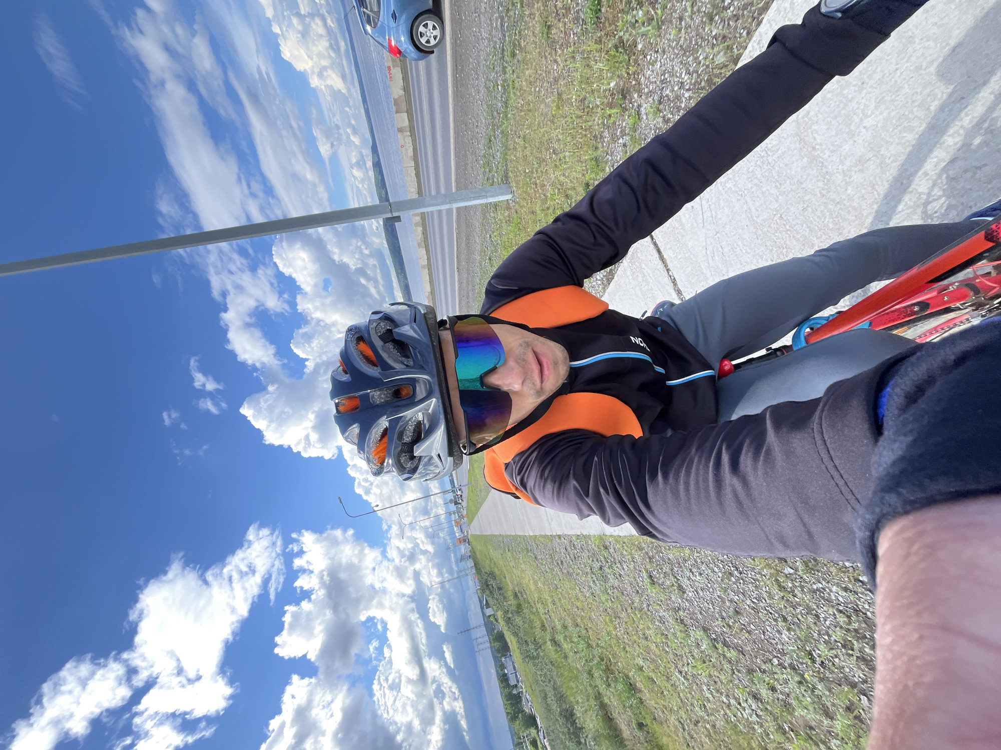 This is a vibrant daytime selfie captured from the perspective of a cyclist in Chaikovsky, Russia. The main subject is a male, looking directly at the camera, wearing a grey cycling helmet, reflective blue and purple sunglasses, a black long-sleeved cycling top with orange shoulders, and grey cycling pants. A partial view of his right arm, clad in a black long-sleeved garment, is in the foreground, suggesting he is holding the camera or handlebars. Part of a red bicycle frame is visible in the lower right.
The setting is outdoors, alongside a concrete sidewalk and a paved road where a blue car is visible in the distance. Beyond the road, a large body of water, likely a river or lake, stretches into the horizon under a vast, bright blue sky filled with scattered white cumulus clouds. Green grass and gravel line the edge of the path. Streetlights are visible along the road in the mid-ground. The weather is clear and sunny, indicative of good conditions for cycling. There is some text on the cyclist's shirt, partially visible as 'NOR' or similar.