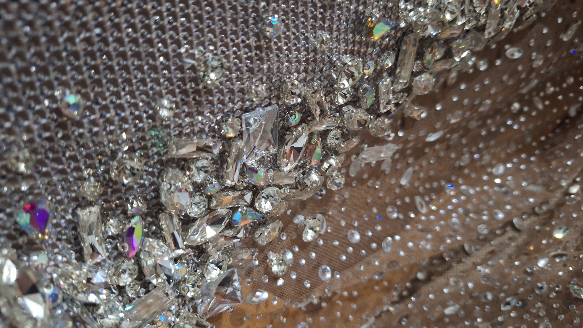 The image shows a close-up of fabric heavily adorned with clear crystals and rhinestones. The fabric on the upper left appears to be a silver mesh, while the rest is a sheer nude color. The crystals are of various sizes and shapes, with some being larger and rectangular, and others smaller and round. Many of the crystals reflect iridescent colors. The background is blurry and out of focus, and there is no information regarding the location being Blagoevgrad, Bulgaria.