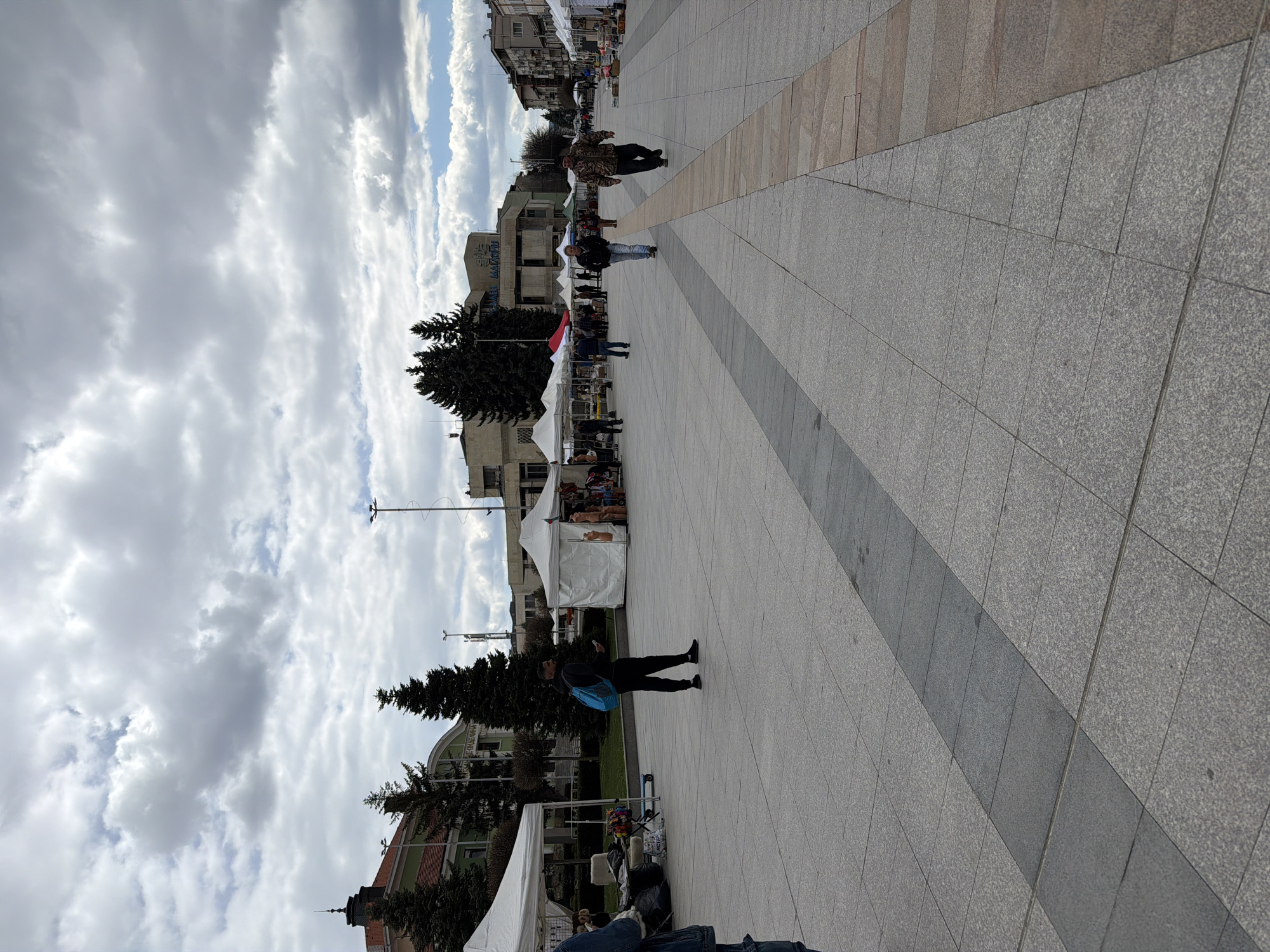 The image captures a street scene in Panagyurishte, Bulgaria. The ground is covered with grey and tan square tiles. People are walking around, with some looking at temporary market stalls set up in the middle of the street. Trees are planted along the street and buildings can be seen on both sides. The sky is cloudy with patches of blue. The image is taken from a high angle.