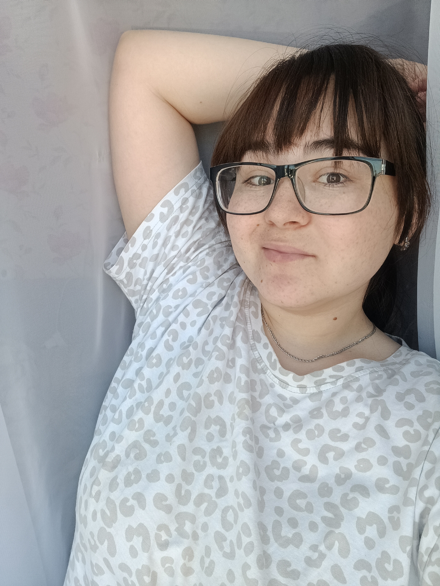 A single individual, a young adult female, is depicted in a head-and-shoulders portrait within an indoor setting in Noviy, Russia. She possesses dark brown hair with bangs, black-framed rectangular glasses, and a light complexion with visible freckles across her nose and cheeks. Her right arm is bent with the hand positioned behind her head or upper neck. She wears a light-colored (off-white or light grey) t-shirt featuring a subtle light grey leopard print pattern and a thin, silver-colored chain necklace. A small, silver-colored earring is present in her right ear. She looks directly at the camera with a neutral facial expression, exhibiting a slight upturn at the corners of her mouth. The background consists of a plain, light-colored fabric, likely a curtain or sheet, displaying vertical folds.
