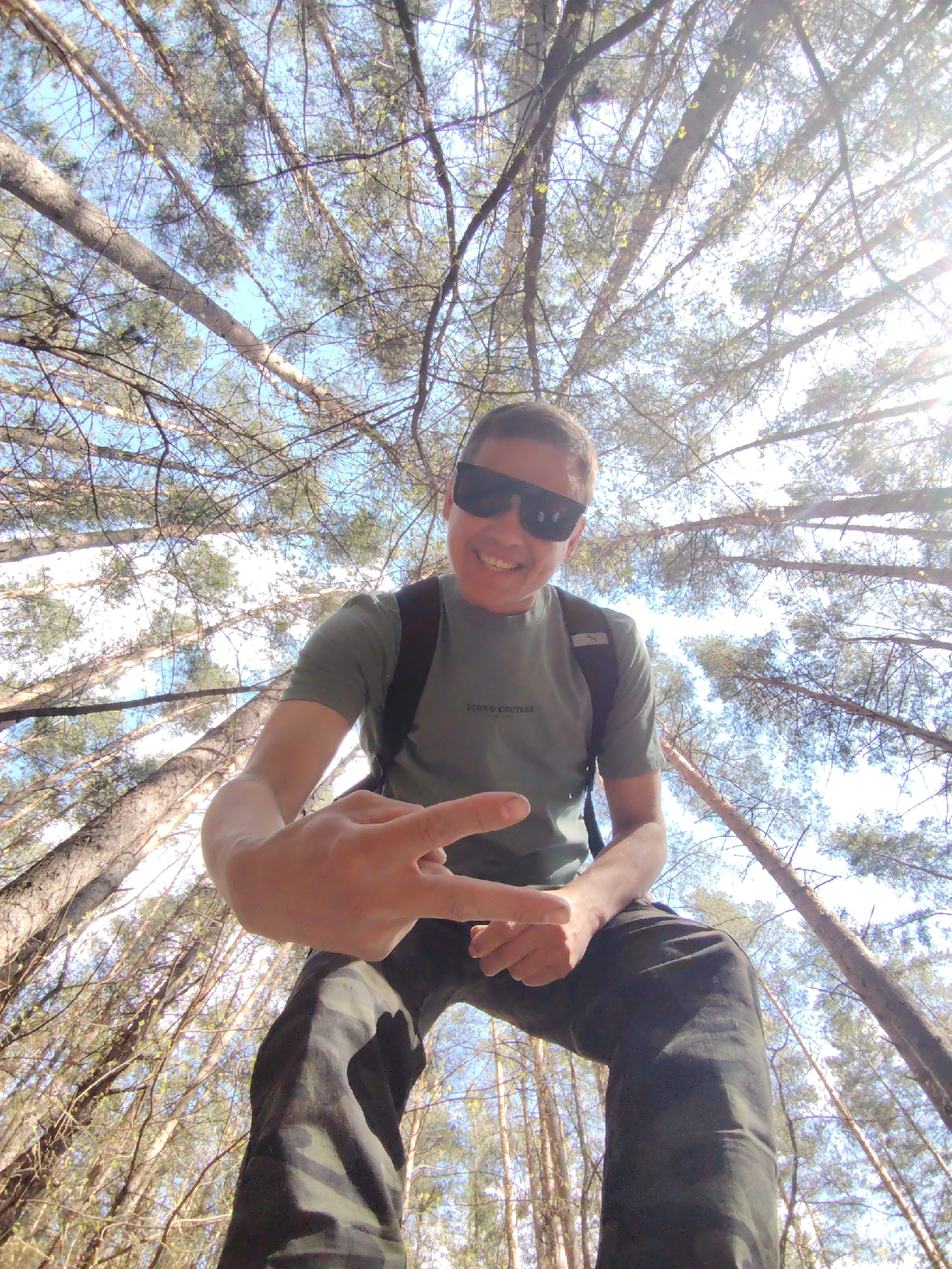 A man stands smiling and making a peace sign in a forest, possibly in the city of Chaikovsky, Russia. He wears sunglasses, a green t-shirt with a logo visible, and camouflage pants. A black backpack strap is visible on his shoulder. The camera is positioned at a low angle, looking up at the man, with a view of tall trees and a blue sky with sunlight.