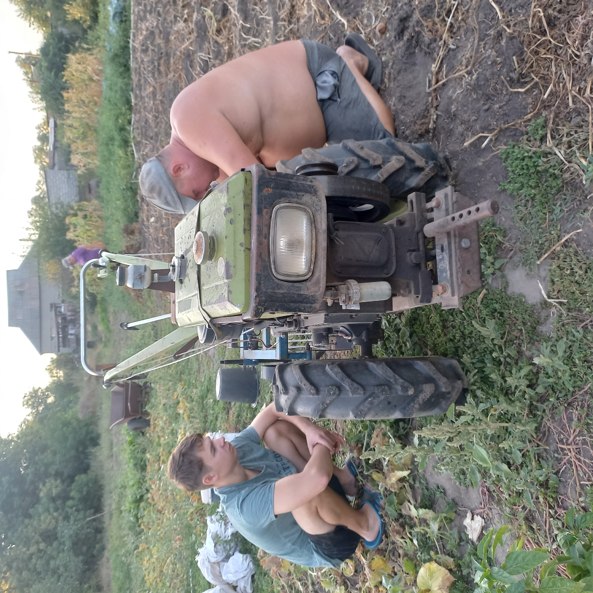 In Klyntsi, Ukraine, two individuals are engaged with a walk-behind tractor in a cultivated field. The image, captured outdoors during daylight, depicts a young man in a teal t-shirt and black shorts, wearing blue flip-flops, crouched near the left rear wheel of the tractor. He is looking to his right, with his arms resting on his knees. To the right of the tractor, a man wearing a grey baseball cap and dark shorts is kneeling. He appears to be working on the front of the tractor, with his head bowed. The walk-behind tractor is green with black accents and features large tires. The surrounding environment consists of tilled soil, low-lying green vegetation, and some dried plant debris. In the background, a building with a pitched roof is visible, along with trees and other structures, suggesting a rural or semi-rural setting. A wheelbarrow and various sacks are also present in the scene, indicating agricultural activity.
