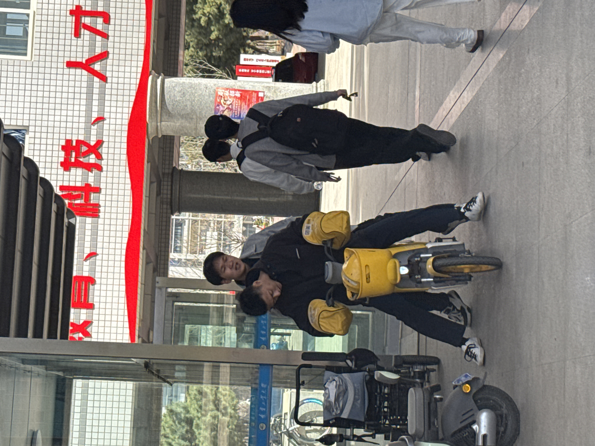The image shows a street scene in Zhongtun, China. Two people are riding on a yellow electric scooter with storage compartments. One person is operating the scooter, and the other is a passenger. There are other people walking on the sidewalk nearby. A parked scooter is visible in the foreground. The building in the background has large red Chinese characters written on its side.