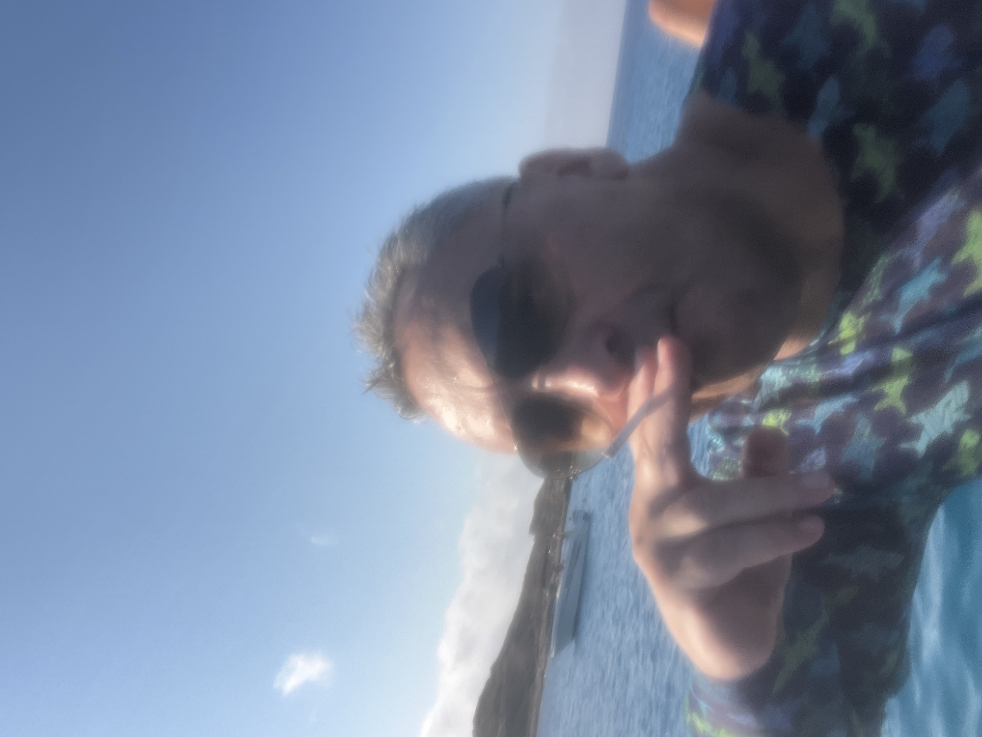 A close-up, slightly blurred, selfie-style photograph captures a man in his late 40s or 50s with graying hair and sunglasses, holding a cigarette between his fingers near his mouth. He is wearing a dark shirt with a repeating pattern of green and blue shapes. The background suggests a sunny day at sea, with a clear blue sky and a body of water. In the distance, a small boat is visible on the water, and a hilly, sparsely vegetated coastline can be seen. The image appears to have been taken in the vicinity of Neo Chorio, Cyprus, a location known for its coastal scenery and clear waters.