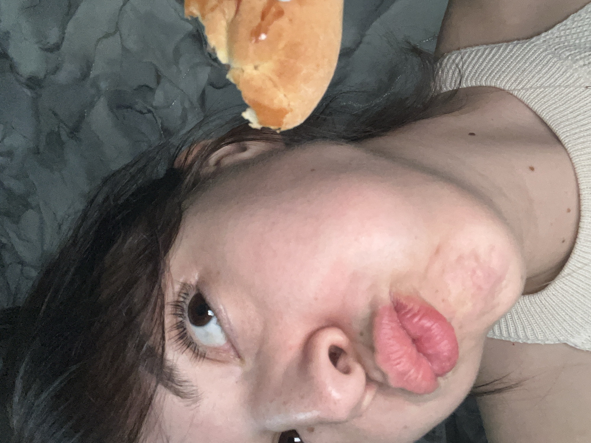 A close-up, skewed-angle view of a person with dark hair, puckered lips, and an upward gaze, a partially eaten pastry is positioned above their forehead. The background includes a textured gray surface and a portion of a cream-colored garment on the right side of the frame. There is no information that would help in identifying the location in Shushary, Russia.