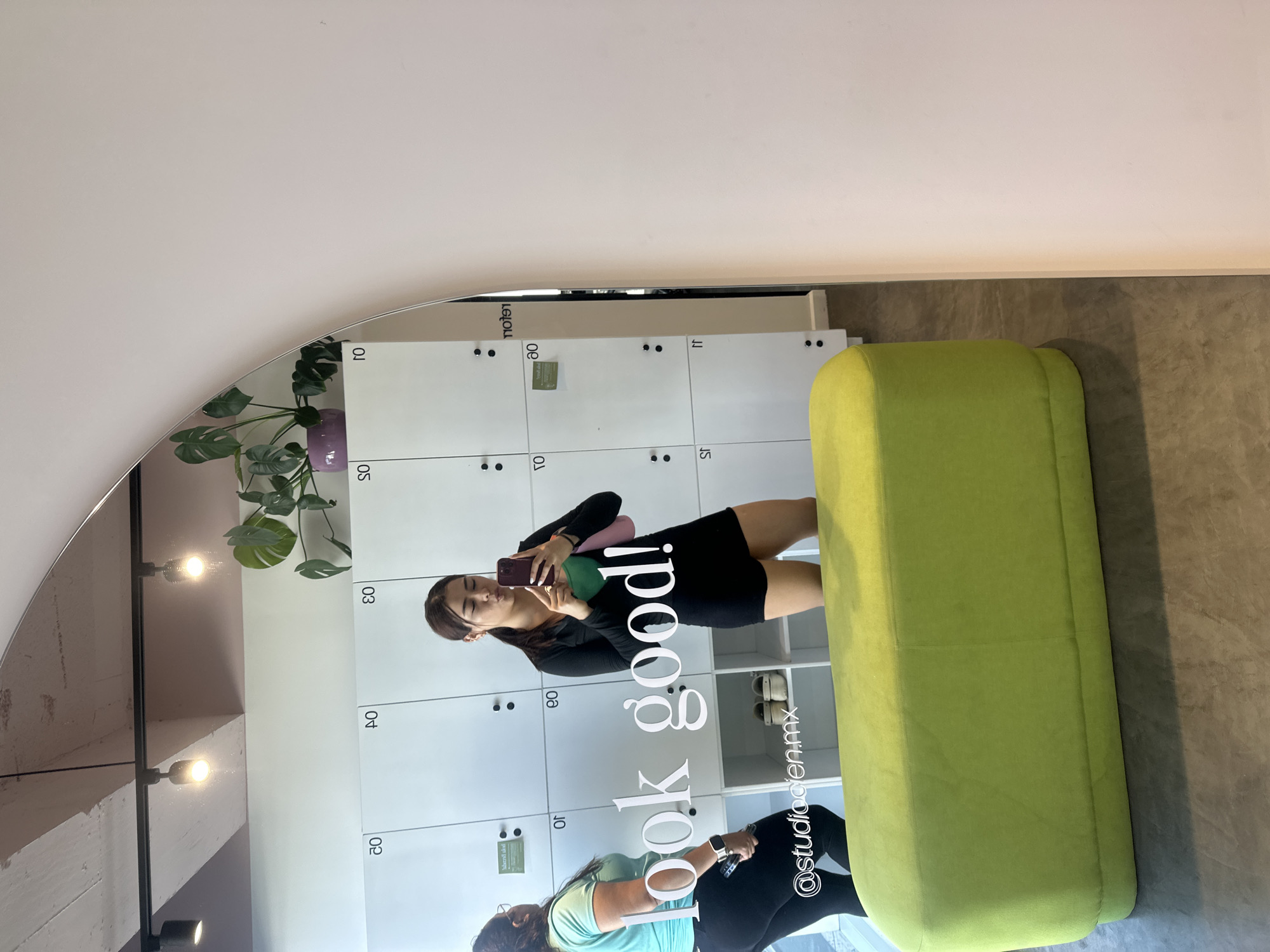 A mirror reflection shows a room at the @studiocien.mx in Puebla, Mexico. In the foreground is a lime green upholstered bench. The reflection shows a young woman, with her hair pulled back, holding a cell phone and a pink and green object to her face. She is wearing a black dress or top and skirt. Behind her are several white lockers numbered 01, 02, 03, 04, 05, 07, 08, 09, 10, 11. On a shelf behind the lockers are white shoes. At the bottom of the lockers, in the reflection, a second woman with dark hair in a ponytail and a light blue top is holding an object in her hand and looking down. On top of the lockers is a green plant in a purple pot. Track lighting runs across the ceiling.