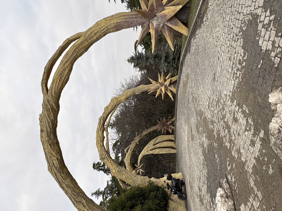 The image displays the entrance to Mtatsminda Park in Tbilisi, Georgia. The scene features multiple large, golden-yellow architectural arches, sculpted to resemble massive, textured tree branches or roots. These arches span a cobblestone paved driveway. At the base of several arches are prominent, multi-pointed, starburst-shaped sculptures, also rendered in a golden-yellow color.
A black scooter, equipped with a rear storage box, is parked on the left side of the paved driveway. The cobblestone surface exhibits areas of wetness and dark staining, with deposits of dirty white material, consistent with residual snow or ice, present in the lower left foreground. A dark metal gate is visible beneath one of the central arches.
The background consists of dense vegetation, including dark green evergreen trees and bare deciduous trees. The sky is overcast and light grey. No individuals are present in the image.