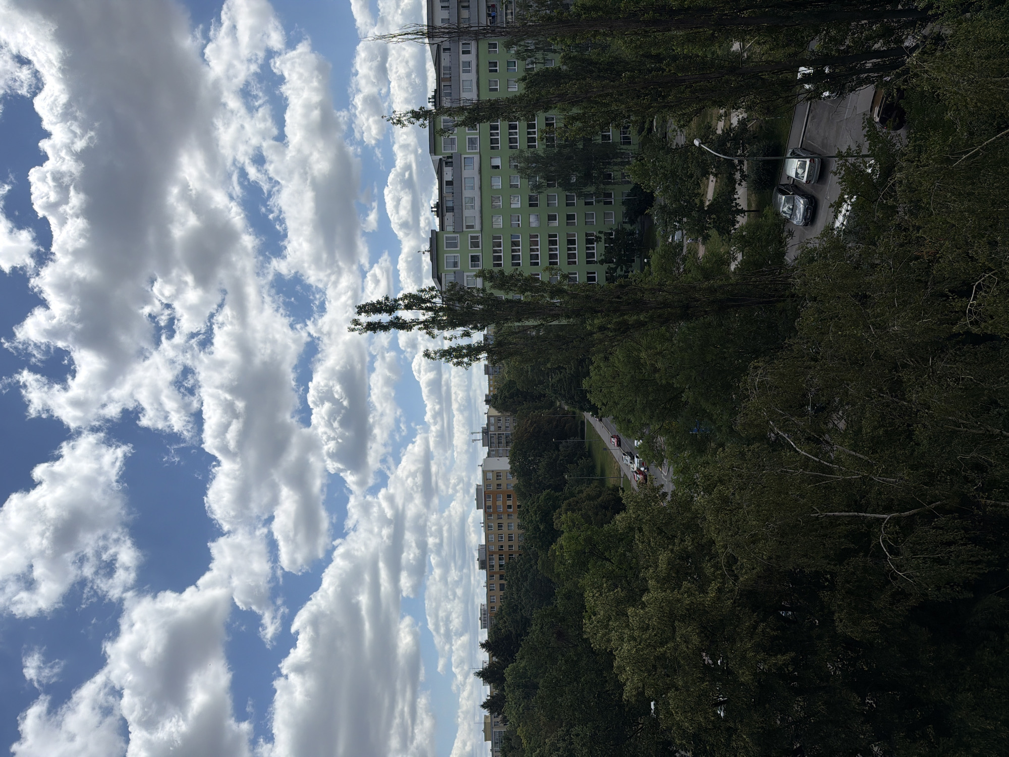 This is an eye-level, long shot, exterior view of a residential area in Prague, Czechia, under a bright, cloudy sky. The image captures a mix of natural and urban elements, with a focus on buildings and greenery. In the foreground, dense, dark green trees dominate the lower portion of the frame, their branches partially obscuring the scene beyond.
Behind the trees, a street runs horizontally across the middle ground, with several cars parked along its side. A few vehicles are also visible driving on the street. Further back, a vibrant green apartment building stands out, characterized by its numerous windows and a light-colored roofline. The building appears to be several stories high and is situated amongst more trees and other structures.
In the background, a row of multi-story residential buildings, varying in color from yellow to light gray, stretches across the horizon. These buildings also feature many windows and are topped with various rooftop structures, possibly including antennas. The sky above is a brilliant blue, dotted with numerous large, fluffy white clouds that cast subtle shadows. The overall impression is one of a suburban or urban residential neighborhood on a clear, possibly warm day.
