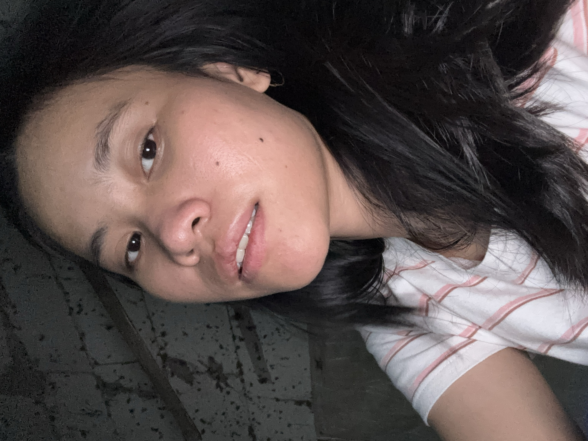 The image is a close-up selfie of a young woman with dark hair and light skin. She is wearing a white t-shirt with thin pink stripes. Her face is turned slightly towards the camera, and she has a mole on her right cheek. The background appears to be a dark, textured wall, possibly brick or concrete. The lighting is dim, suggesting an indoor setting. The image does not contain any elements that would allow for the identification of a specific location in East Jakarta, Indonesia.