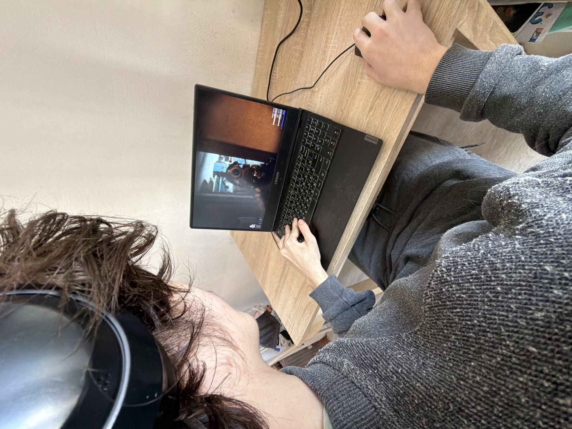 A person is shown from a high-angle perspective, sitting at a wooden desk and using a Lenovo laptop. The laptop screen displays a scene from a first-person perspective video game. The person's left hand is on the keyboard, and their right hand is on a computer mouse. They are wearing a grey sweater and matching sweatpants. The person is also wearing headphones. The location is unknown and cannot be inferred from the image.