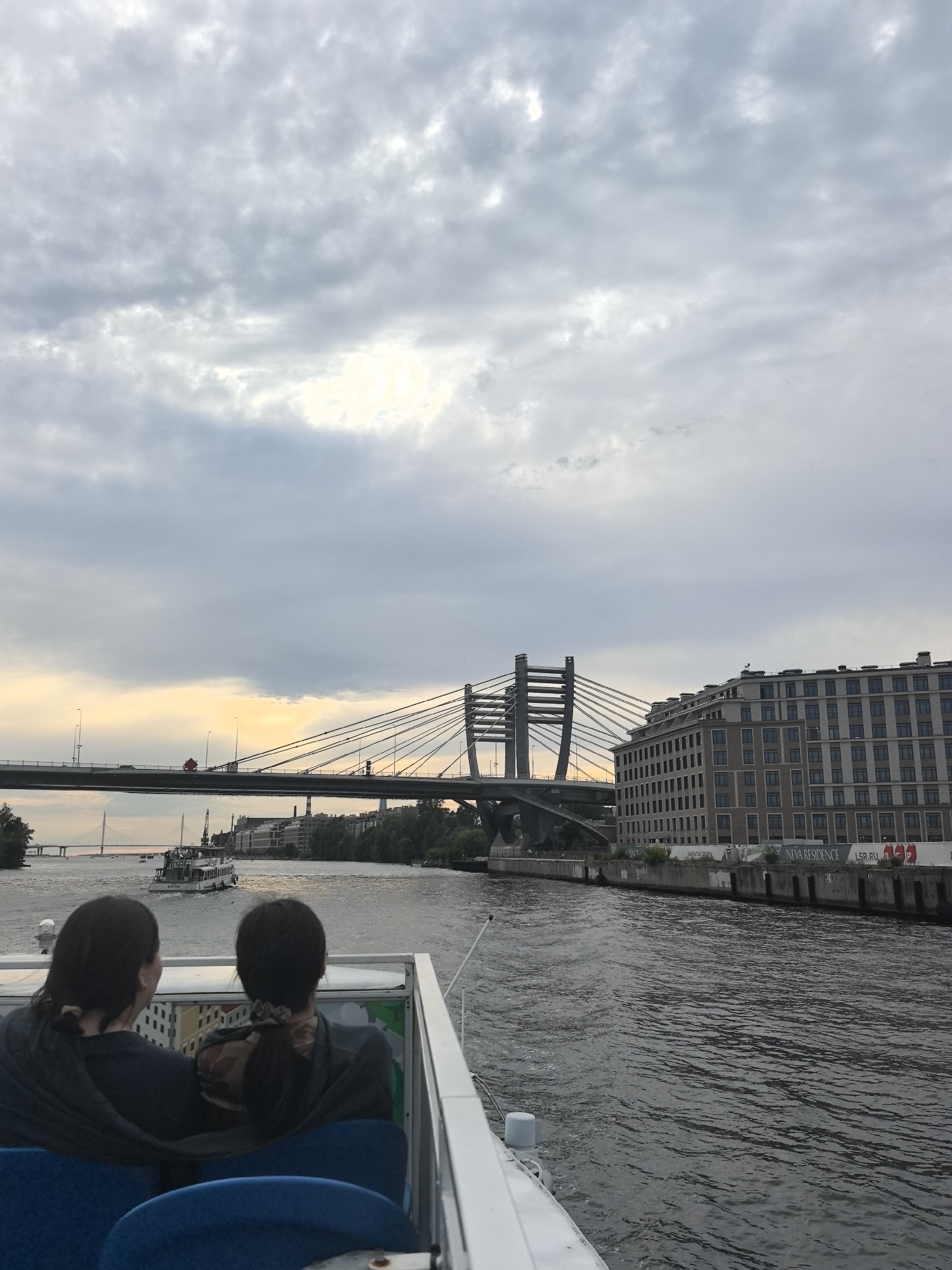 The image depicts a view from a boat traveling on a wide river, likely in Saint Petersburg, Russia, given the architectural style and prominent bridges. In the foreground, the backs of two individuals are visible, seated on blue seats and looking out at the scene. They appear to be on an excursion or tour.
The river is calm, with gentle ripples on the dark water. In the middle ground, a passenger boat is moving away from the viewer. Beyond the river, a large cable-stayed bridge spans the water, with cars visible on its roadway. On the right bank, a modern, multi-story building with many windows stands prominently. On the left bank, there is greenery and hints of other structures, suggesting an urban waterfront. The sky is overcast with dramatic clouds, and a sliver of pale yellow light near the horizon indicates either sunrise or sunset.
Notable visible text includes "NEVA RESIDENCE" and "LSR.RU" on signage along the waterfront, which further confirms the Saint Petersburg location and suggests the presence of a residential development. The overall mood is serene, with a sense of travel and observation. The weather appears to be mild, with the overcast sky creating a diffused light. The time of day is either early morning or late evening.