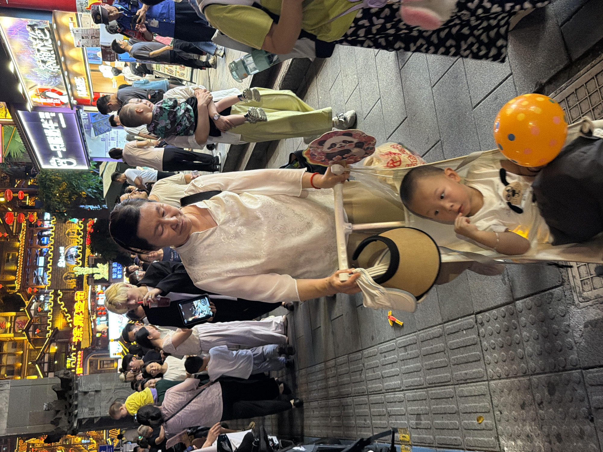 An evening street scene in the Yuzhong District of China captures a crowd of people navigating a paved pedestrian walkway lined with illuminated buildings. In the foreground, a woman pushes a stroller containing a young boy wearing a white t-shirt with a graphic design. The boy is looking to his left, with his finger near his mouth. An orange balloon with blue and orange polka dots is situated beside him in the stroller. To the left of the stroller, a woman in a white shirt holds a beige sun visor and a plastic bag. Behind her, a young boy in a blue shirt walks away from the camera. The background is filled with numerous individuals, some holding drinks, and the facades of buildings adorned with vibrant lights and signage. The overall atmosphere suggests a bustling commercial or entertainment area.