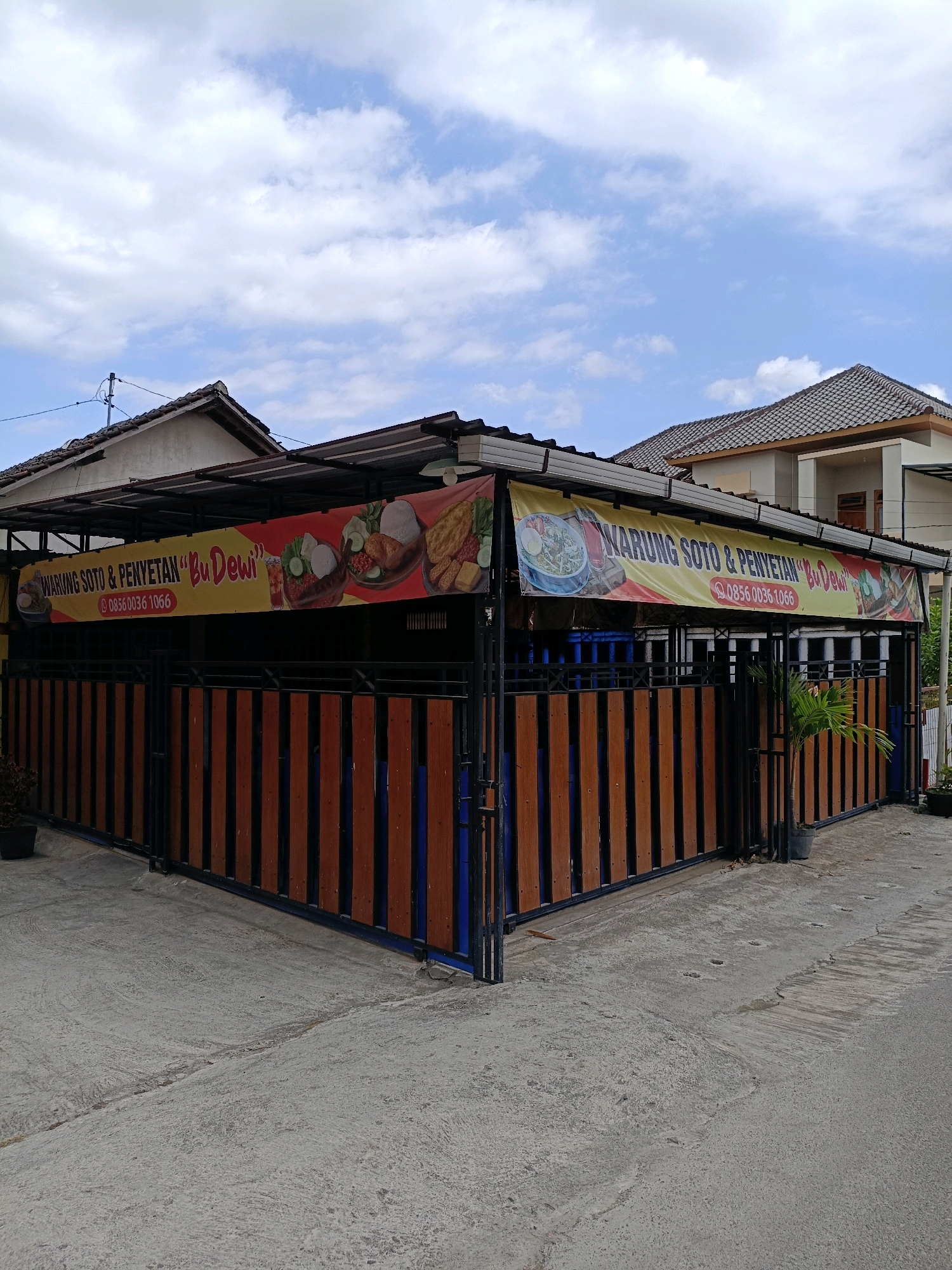 The image shows a street-level view of "Warung Soto & Penyetan Bu Dewi," a food establishment located in Maguwoharjo, Indonesia. The warung features a prominent yellow banner advertising its offerings, with images of various Indonesian dishes such as soto and penyetan. The building has a corrugated metal roof supported by a frame, and a fence constructed with vertical wooden planks and metal accents encloses the area. The ground is paved with concrete, showing some signs of wear and tear. In the background, a two-story house with a tiled roof is visible, suggesting a residential neighborhood. The sky is partly cloudy with a clear blue expanse. There are no people visible in the frame.