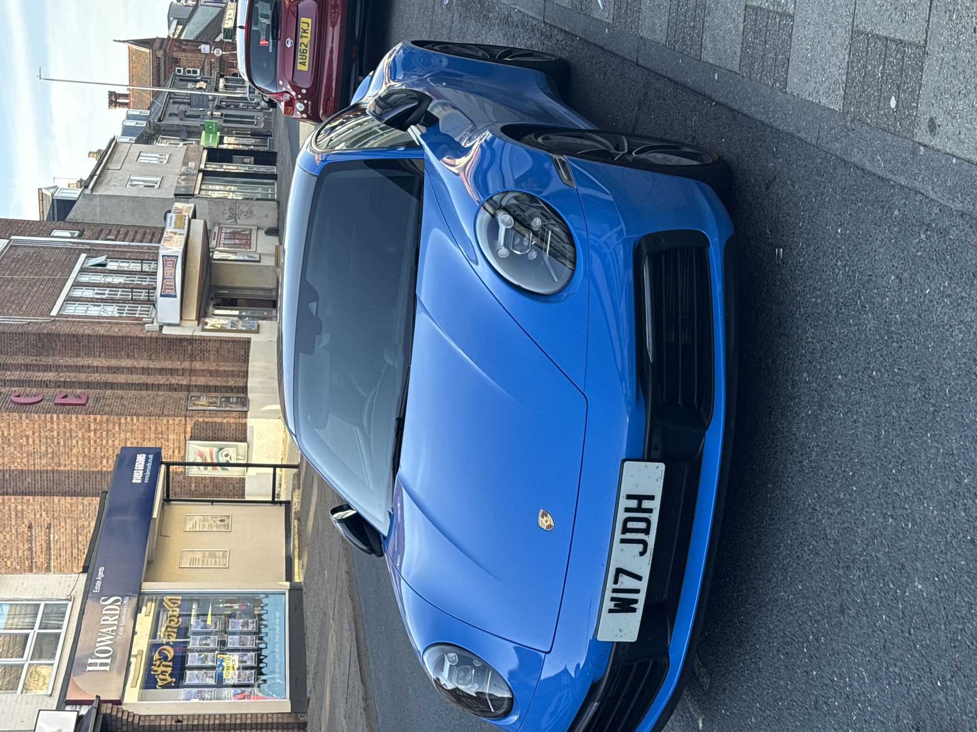 The image shows a bright blue Porsche parked on a street in Great Yarmouth, United Kingdom. The car is positioned along the curb, partially blocking the view of other vehicles parked behind it. The license plate number is visible on the front of the car and appears to be "WI7 JDH". The background reveals a typical street scene with various buildings, including storefronts and residential structures. A building with the sign "Howards Estate Agents" is visible on the left side of the image. The overall setting suggests a commercial or mixed-use area within the city. The image seems to have been taken during daylight hours.