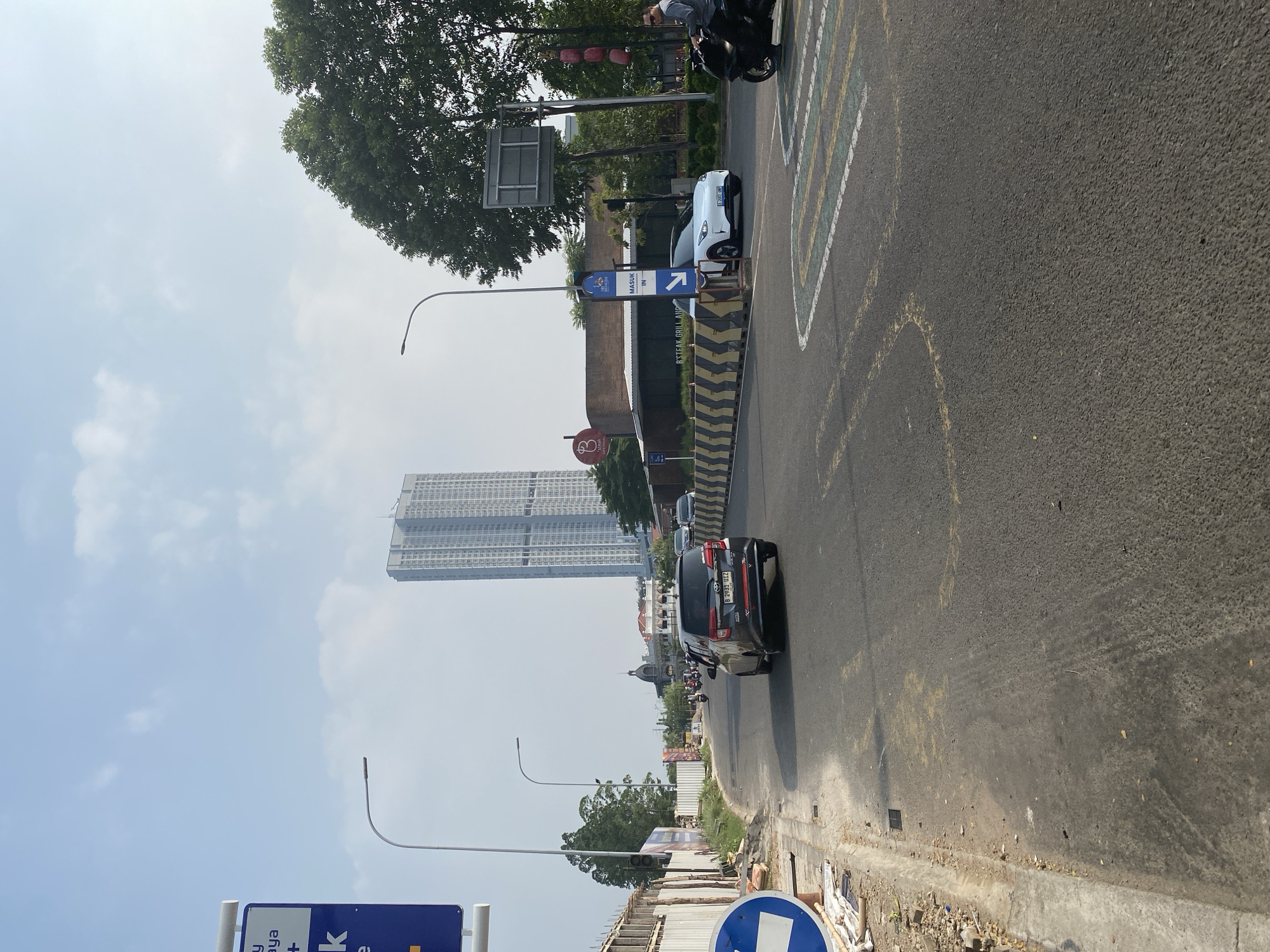 The image depicts a street scene in East Jakarta, Indonesia, on a bright, possibly sunny day given the clear sky and strong shadows. The perspective is from the side of the road, looking forward along the asphalt.
In the foreground, the road surface is visible, marked with yellow and white lines, some of which appear to be part of a traffic lane or a pedestrian crossing. A concrete curb separates the road from a dirt and debris-strewn area, suggesting ongoing construction or maintenance.
Several vehicles are present. A black SUV is prominently featured in the middle ground, driving on the road. Further ahead, a white car is parked near a sign. A black motorcycle is partially visible in the upper right, with a person on it. More motorcycles are seen in the distance, indicating typical traffic in the area.
A tall, modern skyscraper with many windows dominates the background, providing a strong indication of an urban environment. Trees with lush green foliage are also visible, contrasting with the man-made structures. Streetlights are present, suggesting the infrastructure of a developed city.
There is visible text on a blue sign near the white car, which reads "MASUK IN" with an arrow pointing to the right, indicating an entrance. Another blue sign with white text and symbols is partially visible in the lower left corner. A red circular sign with a white symbol is also visible on the left.
The overall scene suggests a bustling urban area with ongoing activity, typical of a daytime scene in a city like Jakarta. The presence of multiple vehicles and the architectural elements point to a well-established commercial or residential district.
