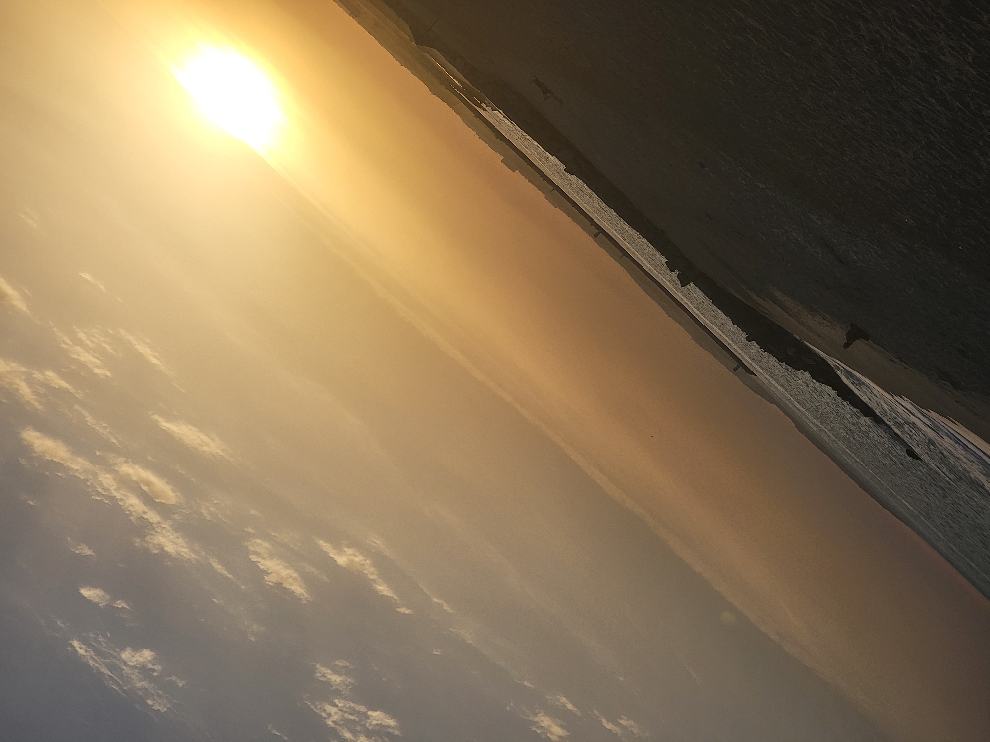The image is a tilted, outdoor photograph of a beach scene at sunset. The sun, a bright orb casting a golden-yellow light, is positioned in the upper right portion of the frame. Wispy clouds are scattered across the sky, transitioning from a pale blue in the upper left to a warm orange glow closer to the sun.
The foreground depicts a sandy beach with subtle wave patterns imprinted on its surface. The water of the ocean meets the beach along the left side of the image, appearing as a dark, textured expanse. A rocky breakwater extends into the sea, separating the water from the sand.
Along the distant coastline, faint outlines of buildings suggest an urban or developed area. A solitary figure appears to be sitting or resting on the sand near the water's edge in the lower right quadrant of the image. The overall impression is one of a serene, end-of-day atmosphere on a coastal location. While the image strongly suggests a beach environment, specific details identifying it as Quarteira, Portugal are not inherently present within the visual information provided.