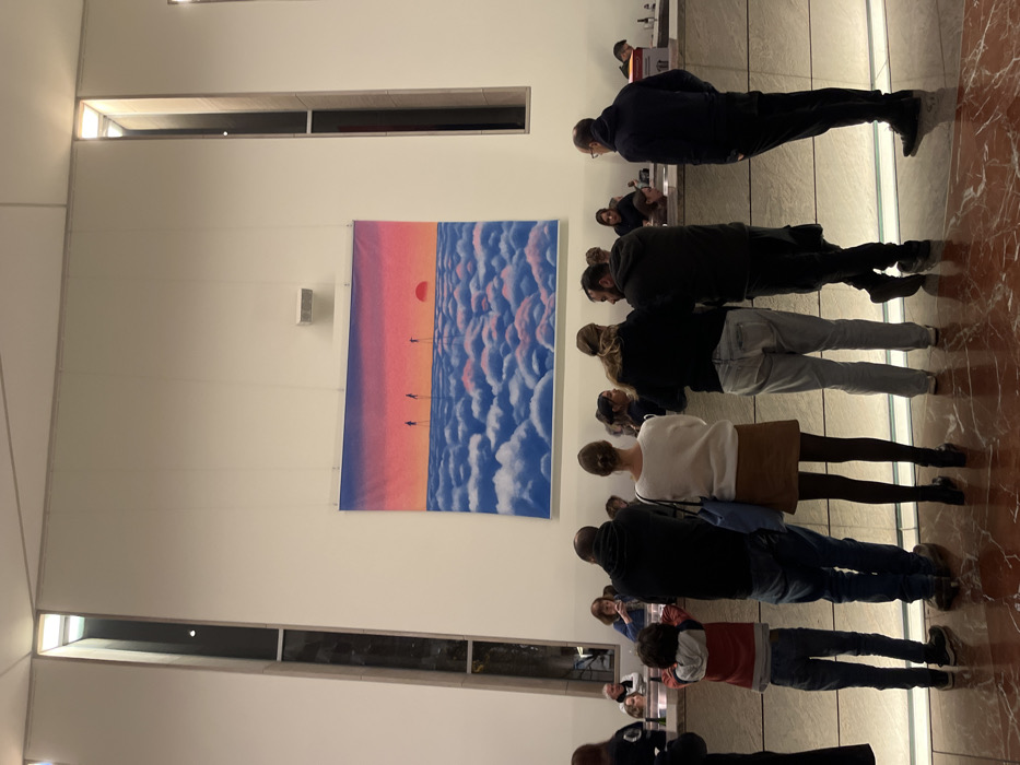 A group of people stand in a room, some looking up at a large art piece hanging on the wall. The artwork is a stylized image of a sunset over clouds. The room has high ceilings, with lights recessed into the ceiling. Long windows are visible on the left side of the frame. The floor is a reflective surface, mirroring the people standing on it.
