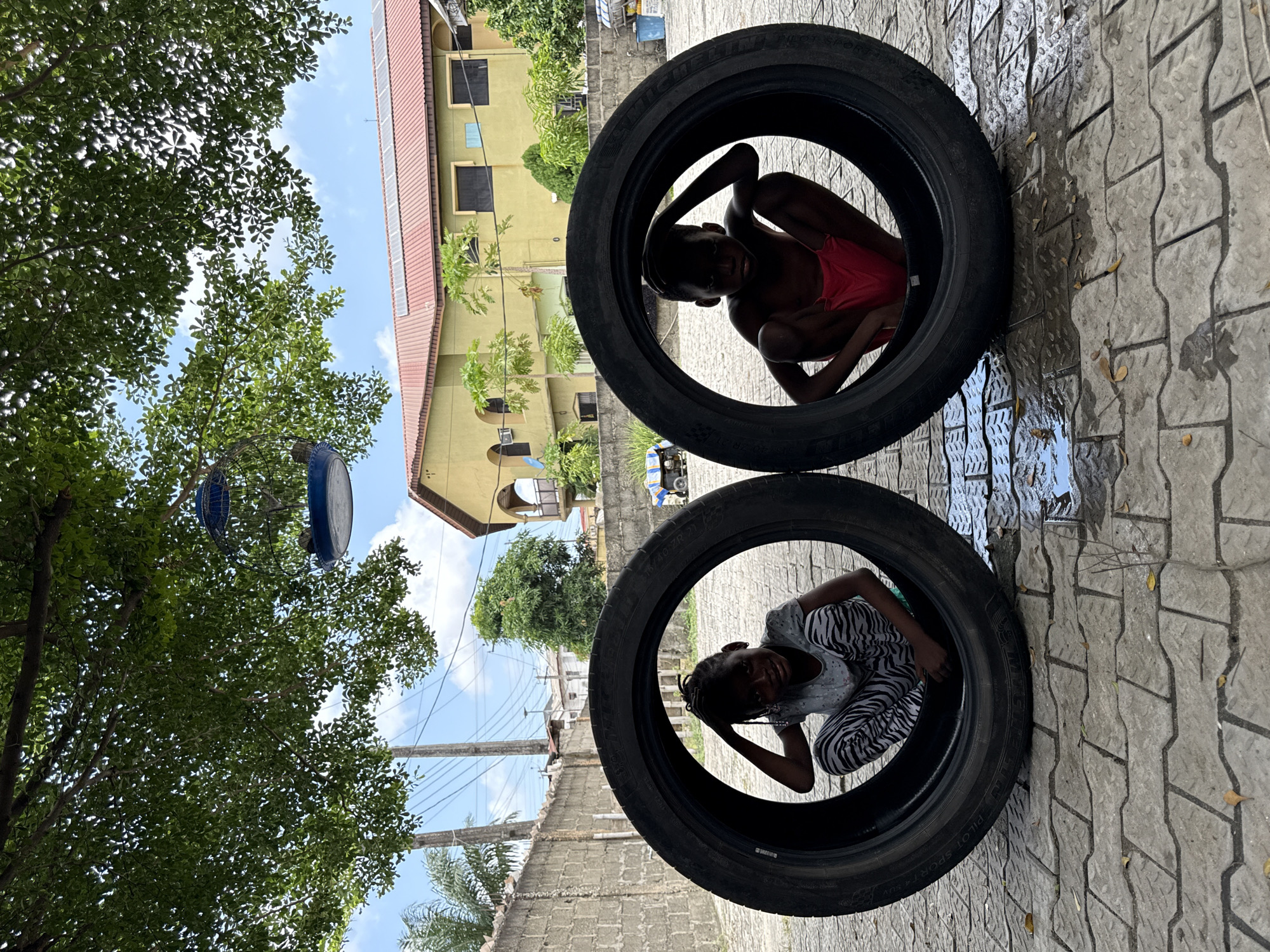 A medium shot depicts two children inside stacked car tires outdoors in Ado, Nigeria. One child is in the top tire wearing a red top, and the other is in the bottom tire wearing a black and white zebra patterned top. Both children have their hands on their heads and appear to be looking at the camera. The tires are stacked on a paved surface that has some water. Behind the tires, there is a concrete block wall and a yellow building with a red roof and green foliage in front of it. In the top left corner of the shot is a tree and the sky. There is an unidentified blue and black object hung in the tree. The image appears to be taken with the camera tilted to the side.