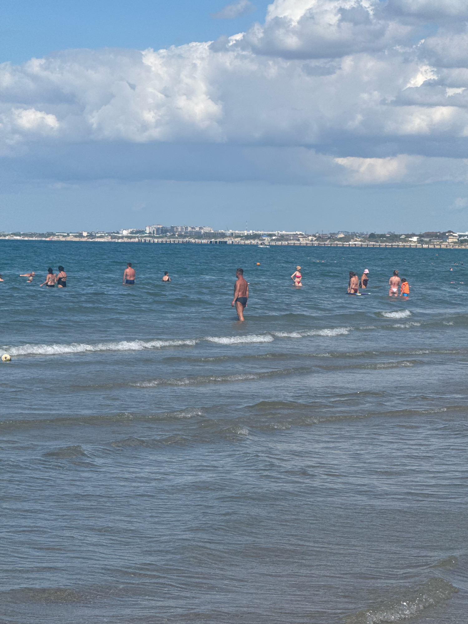 An aerial view captures a serene beach scene in Anapa, Russia, under a partly cloudy sky. The turquoise waters of the sea gently lap against the shore, creating small waves. Several people are enjoying the water, some wading, others swimming. In the distance, a line of buildings forms the Anapa skyline, with a pier extending into the sea. A yellow ball floats near the shore, suggesting recreational activities.