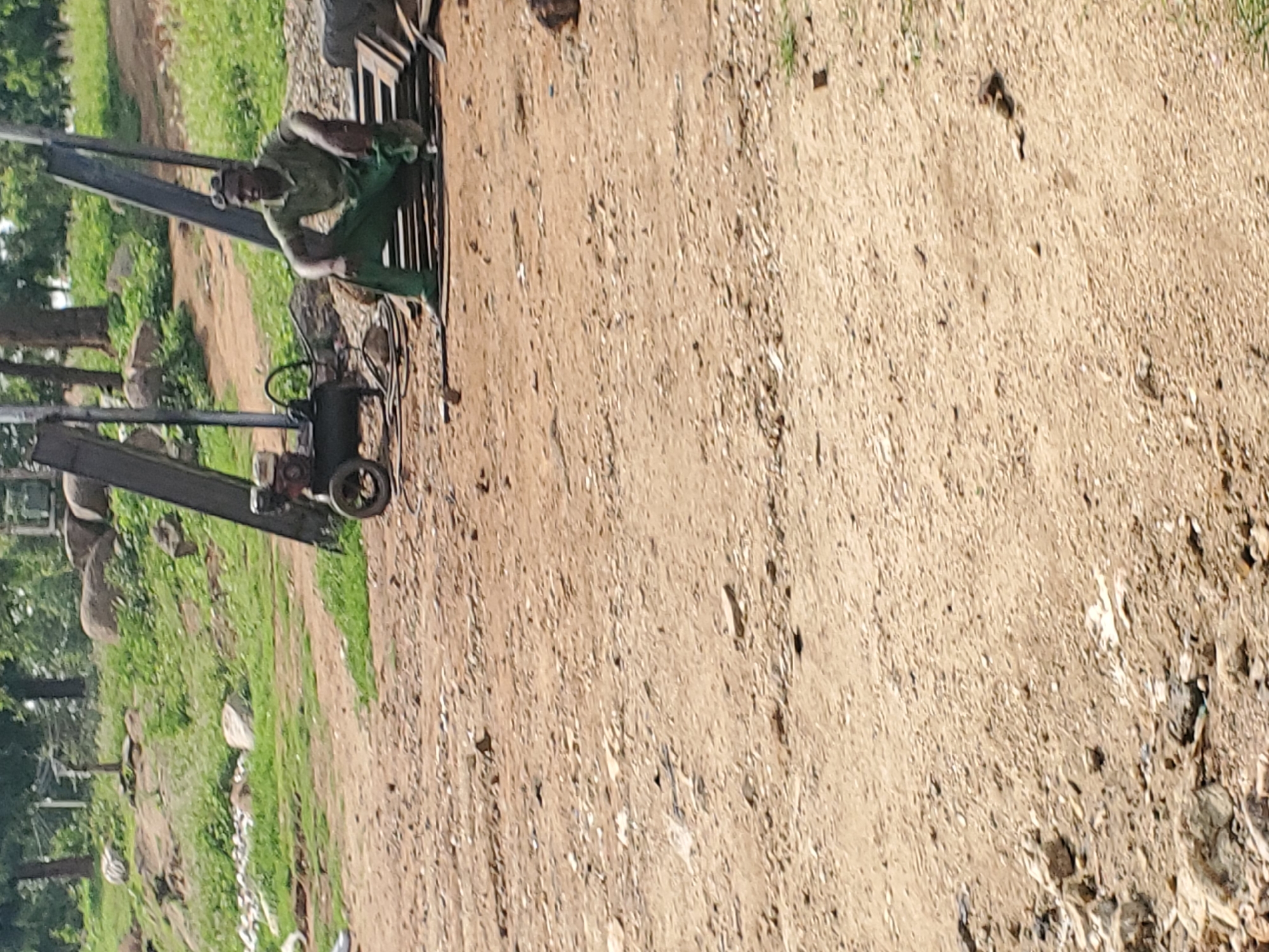 The image shows a person in green work clothes and a hard hat operating a piece of machinery that appears to be a drilling rig. The person is positioned behind the controls, with their hands on a steering wheel-like mechanism. The rig itself is metal and has a large drill bit or auger. In the background, there is a patch of green grass and trees, suggesting an outdoor setting. The ground is made up of dirt and gravel. The scene appears to be a construction site or a similar work area. The time of day seems to be daytime, given the bright lighting. There is no visible text in the image.