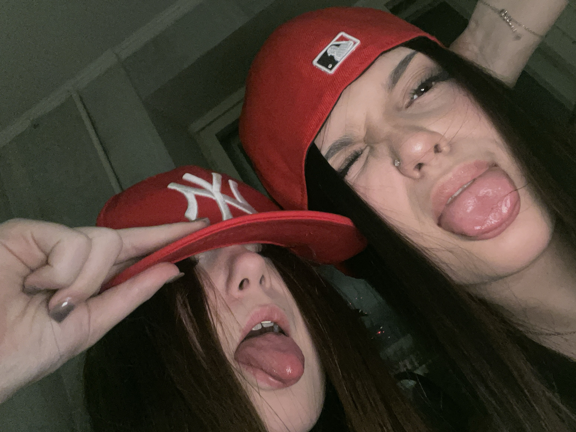 Two young women are posing close to each other, sticking their tongues out for the camera. Both are wearing red baseball caps. The woman closer to the camera has a New York Yankees cap, with the logo partially covered by a hand adjusting the cap. The other woman's cap has the MLB logo. The setting appears to be indoors, though there's not much of the background visible. There is no indication of this image being from Saint Petersburg, Russia.