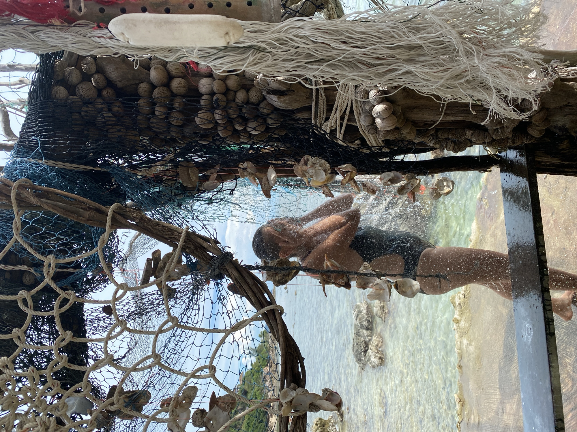 A woman stands in the ocean at ตำบลเกาะพะงัน, Thailand, taking a shower under a makeshift showerhead. She is wearing a black one-piece swimsuit and her body is partially obscured by water droplets. The shower apparatus is constructed from driftwood, fishing nets, shells, and coconuts. The ocean water is clear and shallow, with rocks visible beneath the surface. In the background, a lush green island can be seen. The overall scene evokes a sense of rustic, beachside living.