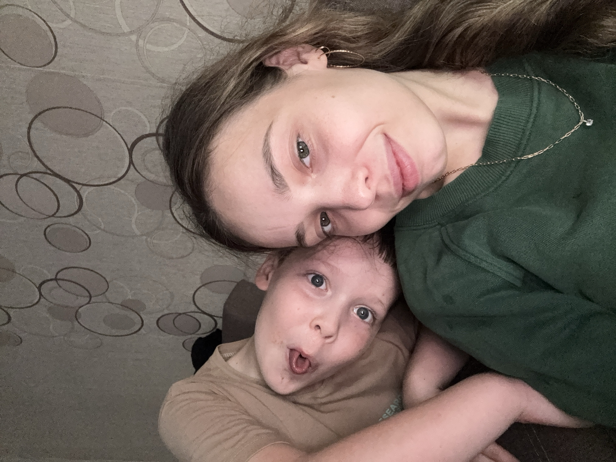 The image shows a selfie taken indoors, likely in a residential setting. A young woman with brown hair, wearing a green sweater and a gold necklace, is in the foreground, smiling at the camera. Her right ear, adorned with a gold hoop earring, is visible. To her left and slightly behind her, a young child with blue eyes and light-colored hair is lying down, looking up at the camera with their tongue sticking out. The child is wearing a tan shirt. The background consists of a textured wall with a circular pattern. The lighting appears to be artificial, casting a soft glow on the subjects. There is no identifiable information within the image to suggest a specific location in the city of Kemul, Russia.