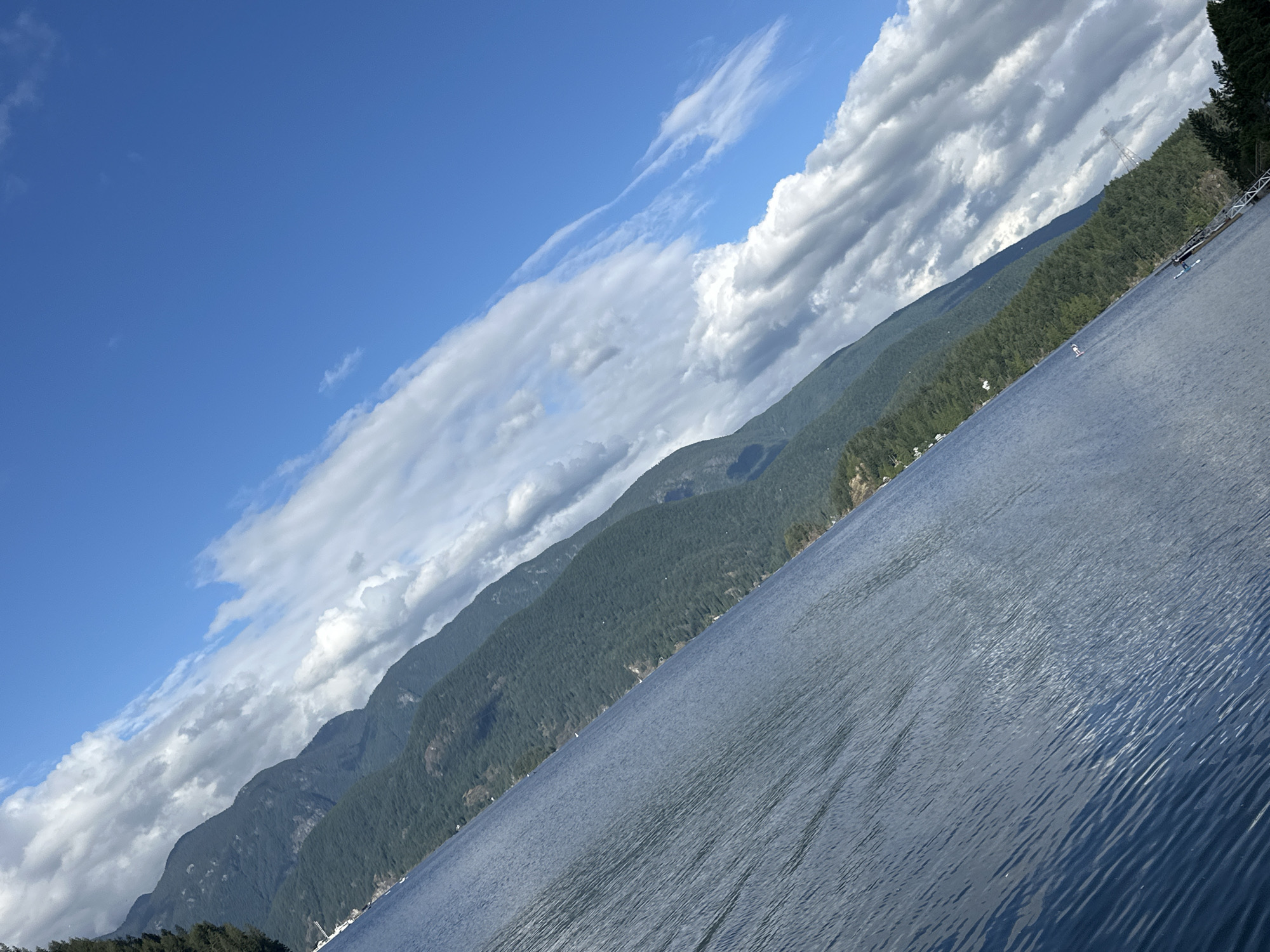The image depicts a panoramic view of a body of water with mountains in the background. The scene is captured from a low angle, likely from a boat on the water.
In the foreground, the water is calm with gentle ripples, reflecting the blue sky. The color of the water is a deep blue, suggesting depth.
The middle ground is dominated by a lush, green mountainside that slopes down towards the water. The vegetation appears dense, consisting of trees and shrubs. Scattered along the base of the mountain, near the water's edge, are several small white structures, likely houses or cabins, indicating a residential area.
The background features a series of imposing mountains, layered and receding into the distance. Their peaks are partially obscured by a dynamic sky filled with a mixture of bright white cumulus clouds and wispy cirrus clouds, interspersed with patches of clear blue sky. The angle of the photograph, with the horizon tilted diagonally, adds a sense of dramatic perspective.
The presence of a pier or dock structure can be observed along the shoreline in the lower right portion of the image, with some small objects that could be boats or watercraft nearby. A tall transmission tower is also visible on the mountainside in the distance.
This scene is consistent with the geography of the District of North Vancouver, Canada, particularly areas along the shores of Indian Arm or Burrard Inlet, known for their mountainous terrain and waterfront communities.