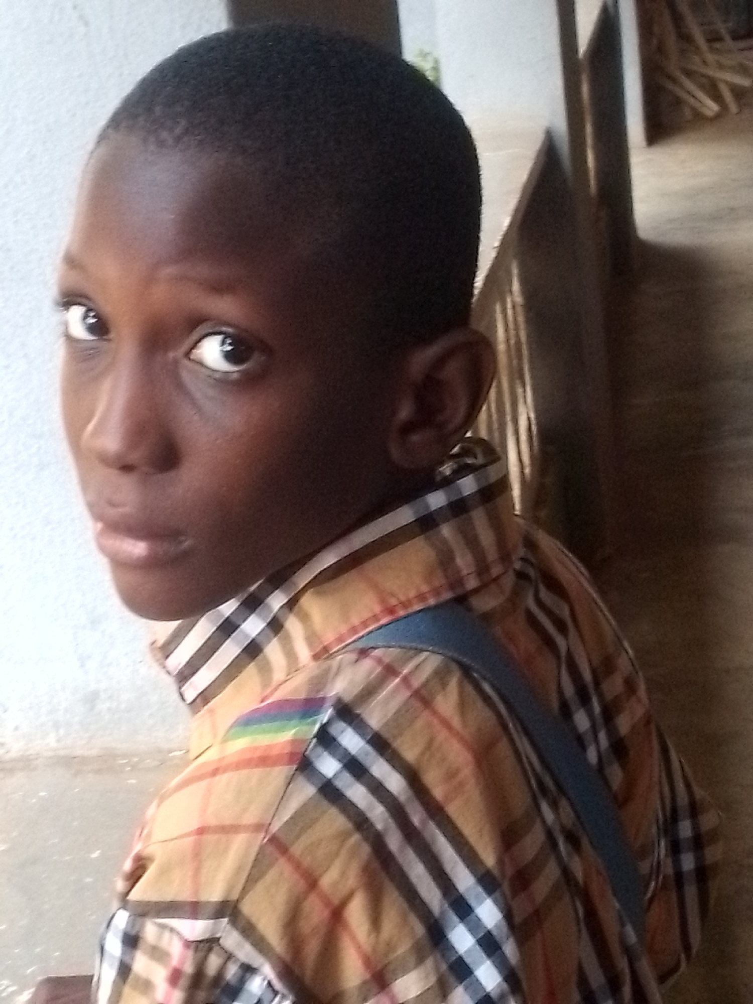 A young, dark-skinned person with close-cropped hair is captured in a medium shot. They are wearing a plaid shirt with a blue strap across their shoulder. The person is looking directly at the camera, and the background appears to be an interior space with light-colored walls. A wooden railing is also visible in the background. The location is Unknown, Unknown.