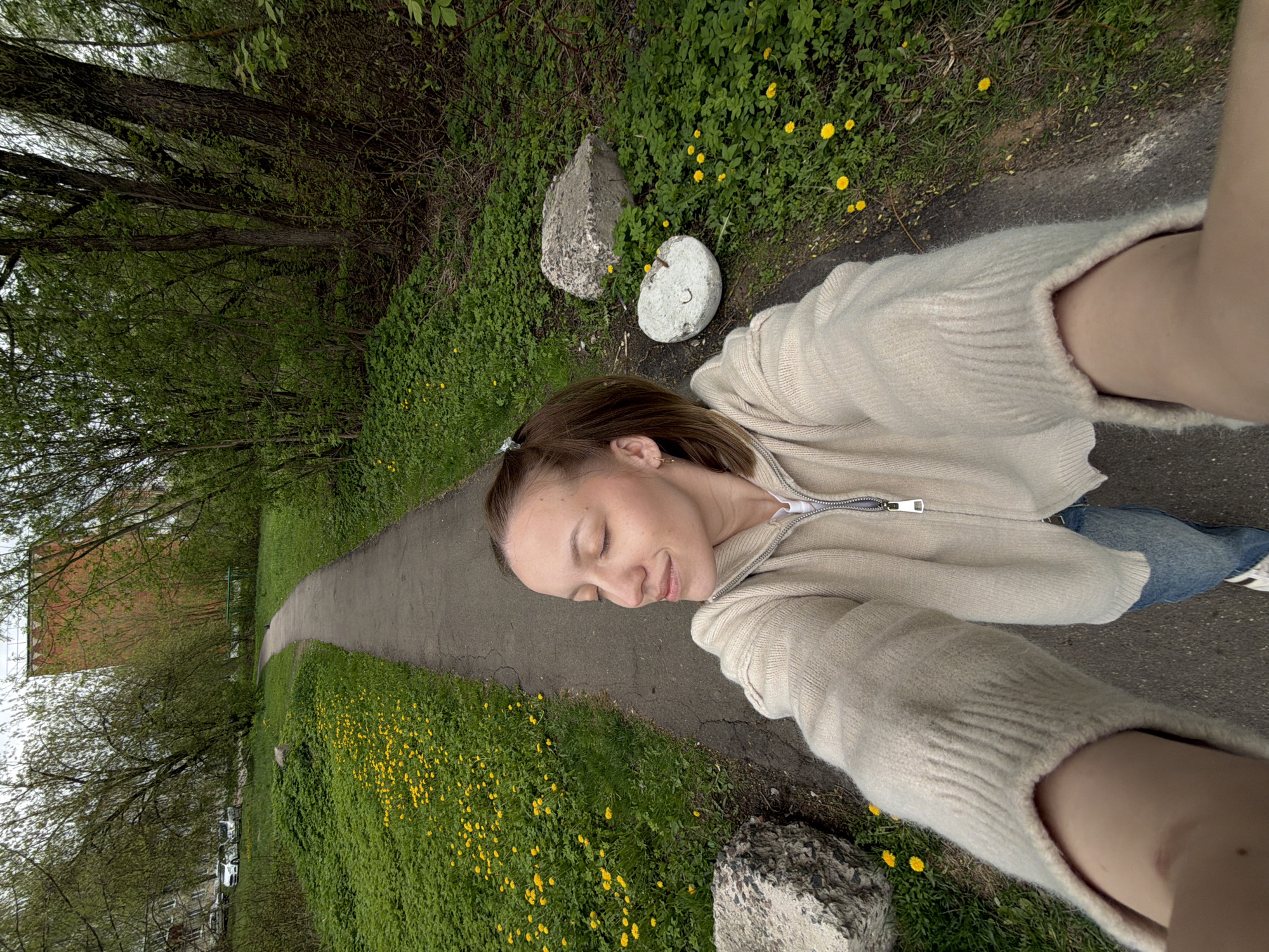 A woman is seen in an unconventional selfie. She is lying on a gray asphalt path in Reshetnikovo, Russia. The path has dandelions growing along its edge and a large building is visible in the background. The woman is wearing a light-colored zip-up sweater and blue jeans. She has her eyes closed and appears to be smiling. There are concrete blocks at various points along the path.