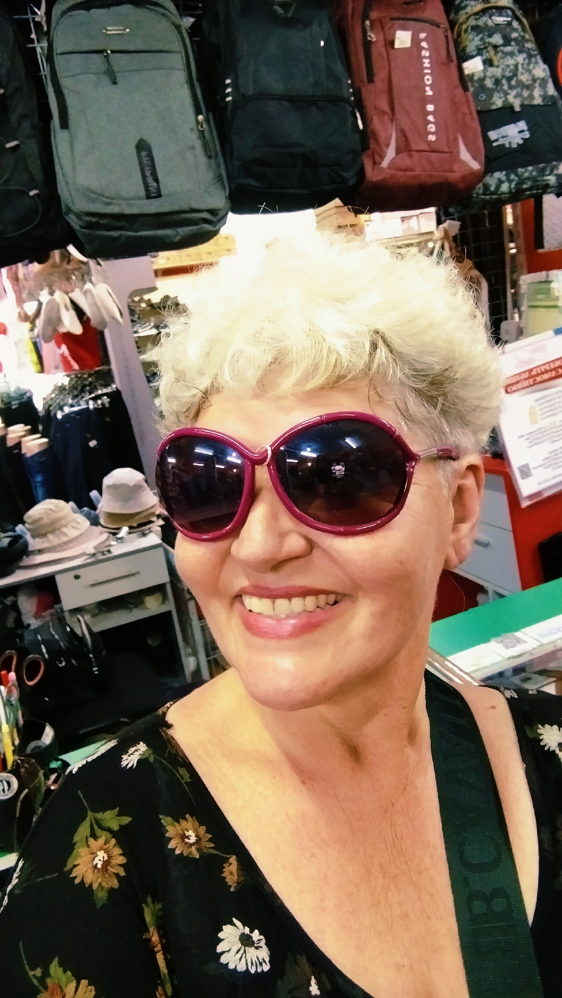 In the city of Horishni Plavni, Ukraine, a middle-aged Caucasian woman with short, white, permed hair smiles at the camera, wearing large, burgundy-rimmed sunglasses. She is wearing a black dress with a floral pattern of white and yellow daisies. Behind her, several backpacks of various colors, including grey, black, and burgundy, are hanging from a rack. To her left, a shelf holds several hats, and a drawer unit is visible. The background also shows what appears to be a retail environment with merchandise and signage.