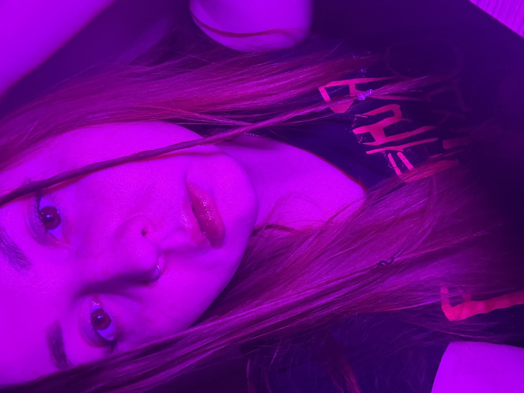 A close-up, tilted shot illuminated by purple light features a person with long, reddish-brown hair. The person's face is visible, including eyes, nose with a ring piercing, and lips. The person is wearing a dark-colored garment, and part of a text design in a bright pink/red color is visible on the garment. A white object, possibly a shoe, is visible at the top of the frame.
There is no information available to determine the location of the scene to be in Izhevsk, Russia.