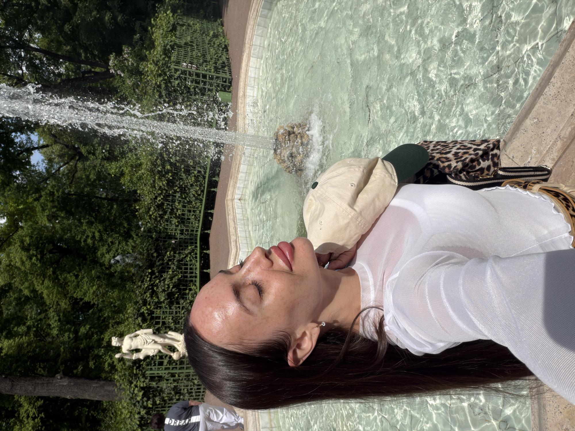 A young woman stands in front of a fountain in a park on a sunny day. She is wearing a sheer white long-sleeved top and has her eyes closed, tilting her head back as if enjoying the sun. She is holding a beige baseball cap over her shoulder, which has a green brim. A leopard-print purse with a striped strap is also visible. In the background, a statue of a man and a woman is visible behind a green fence, along with lush green trees and a tall fountain. The water in the fountain is clear and sparkling. Another person, wearing a gray shirt and white pants, is walking away from the camera in the background. The fountain is circular with a stone edge and the surrounding area appears to be paved with cobblestones. The location is the Summer Garden in Saint Petersburg, Russia.